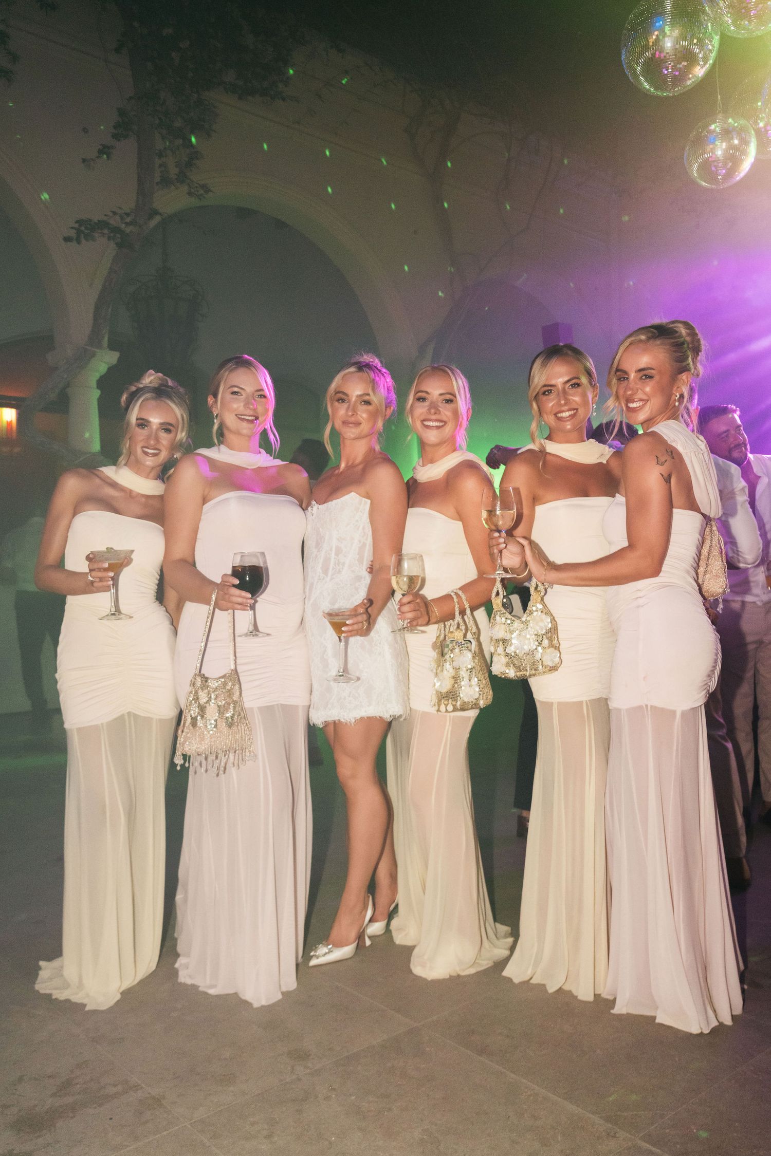 Six bridesmaids in matching white gowns posing together at an outdoor wedding reception party.