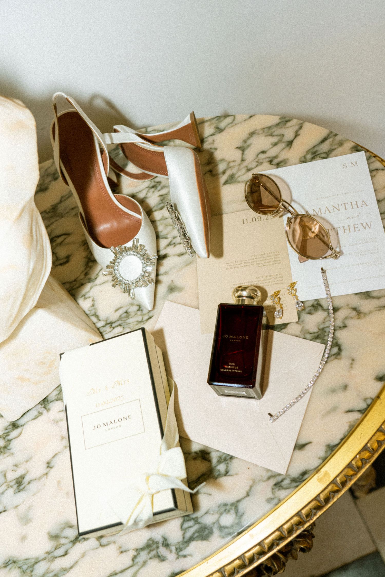 Bridal flat lay on a marble table featuring crystal heels, Jo Malone perfume, sunglasses, and wedding invitation.