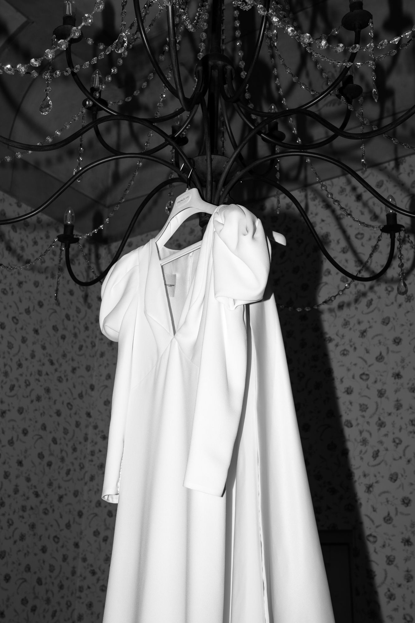 Black and white photo of a elegant white wedding dress with bow detail hanging from a crystal chandelier.