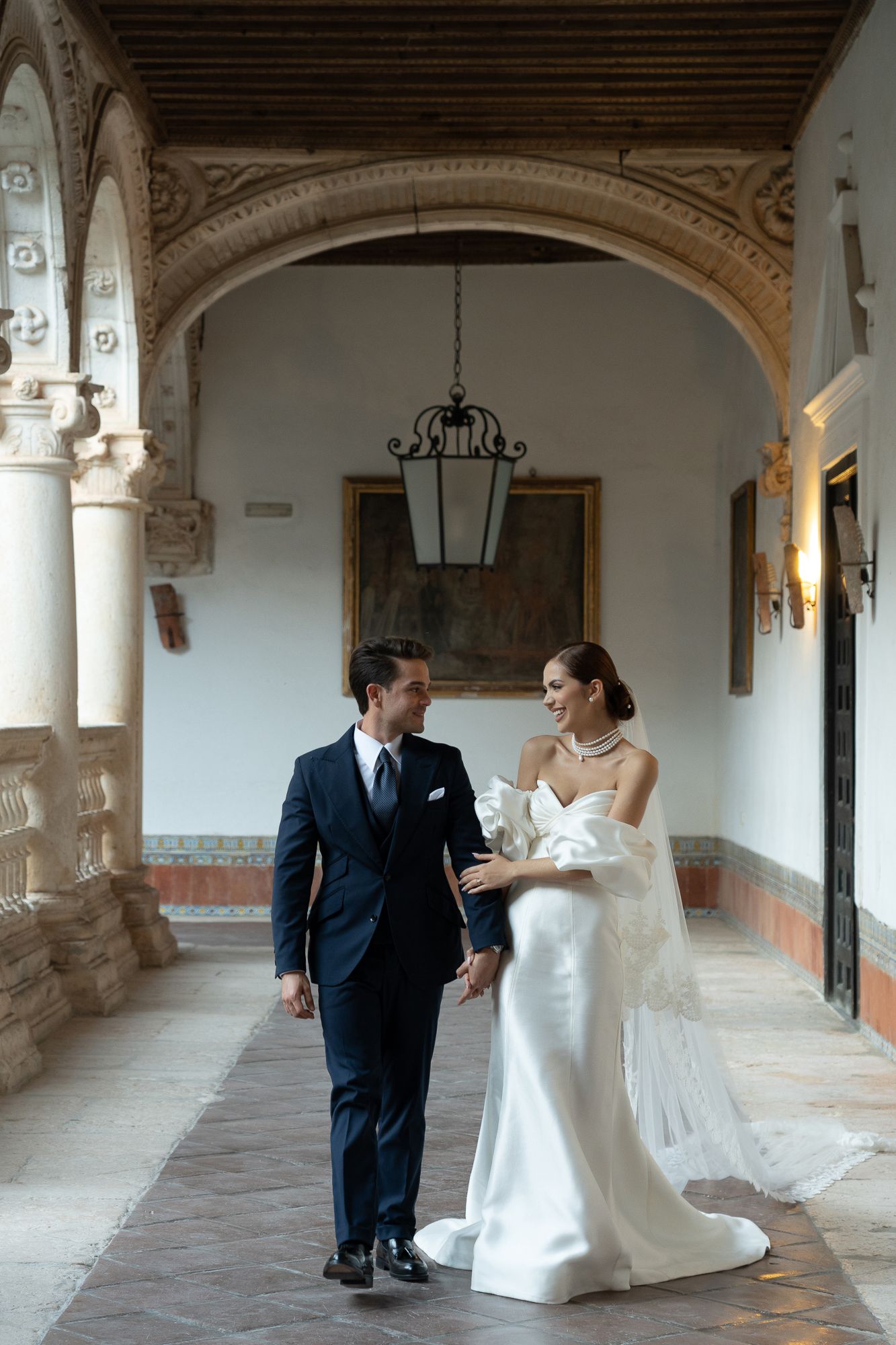 Bride and groom walking hand in hand through elegant arched corridor of historic venue on their wedding day.