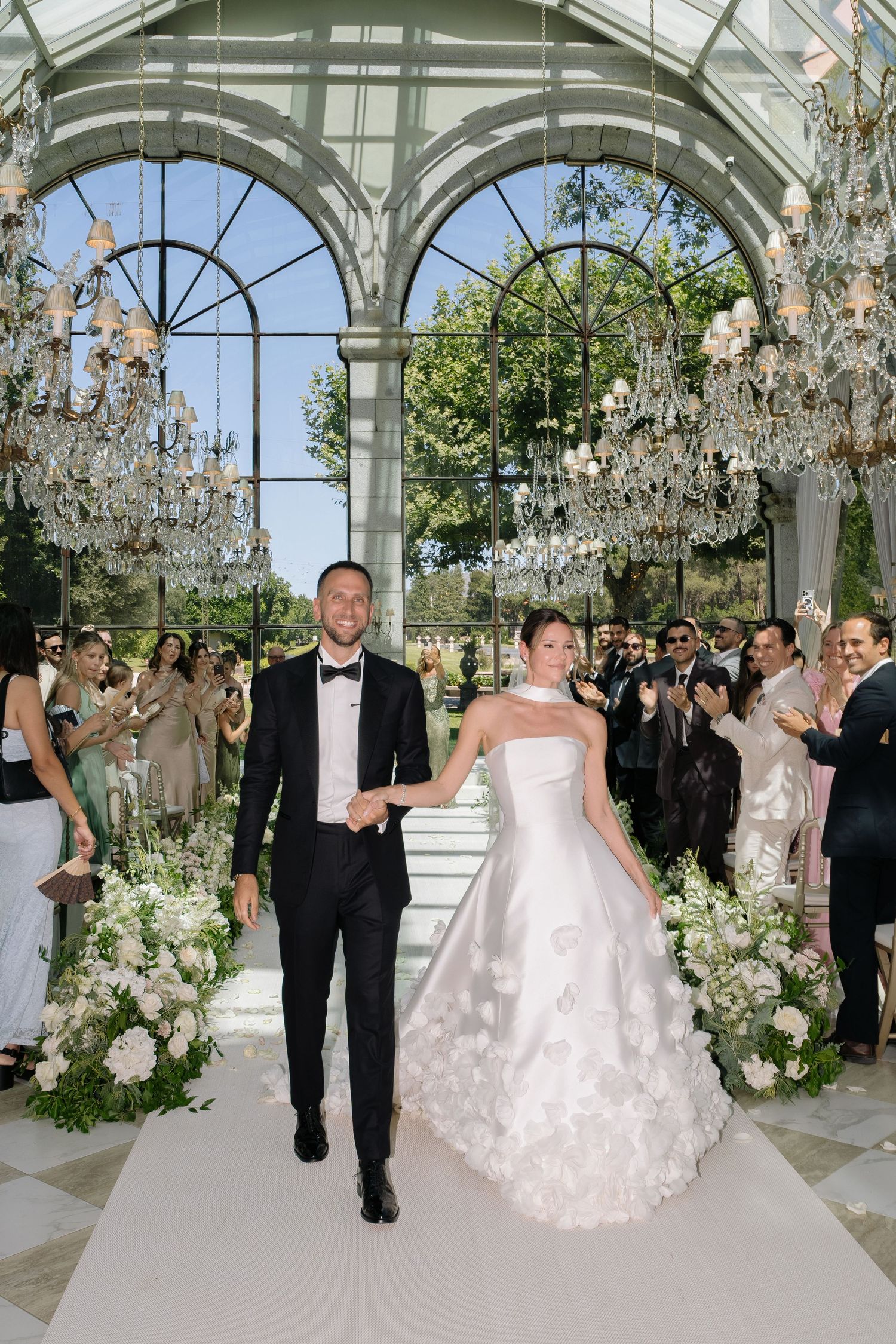 Bride and groom walk down the aisle in an elegant glass conservatory adorned with crystal chandeliers and white floral arrangements.