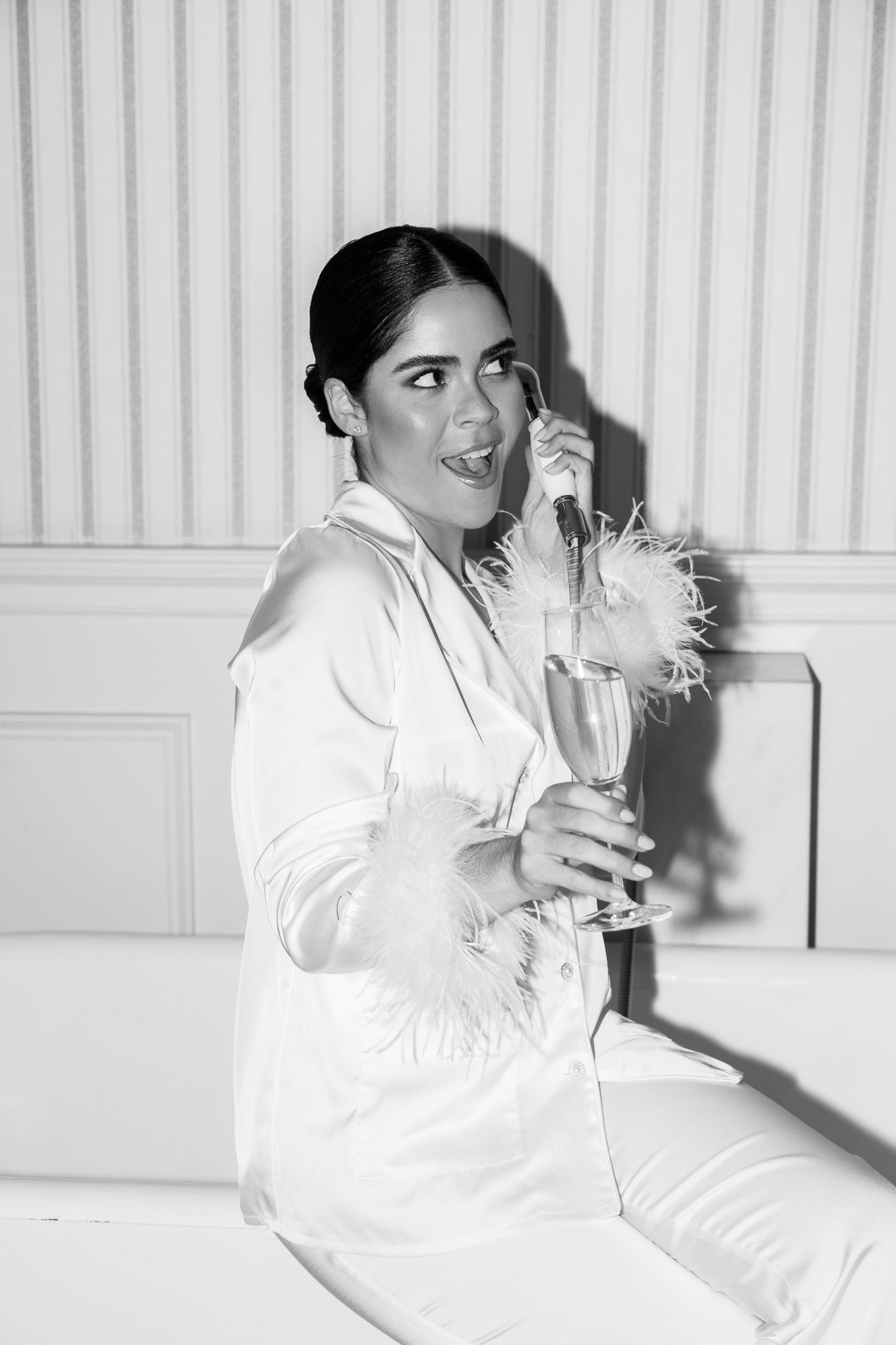 Black and white photo of a woman in white suit holding champagne glass with feather decoration, sitting and smiling.