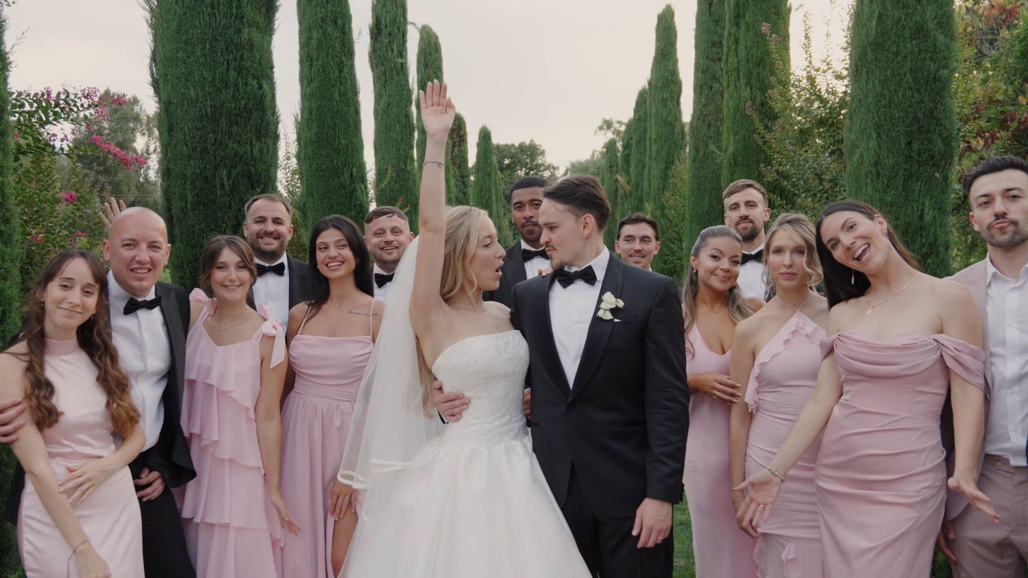 Wedding party poses outdoors with cypress trees, bridesmaids in pink dresses and groomsmen in tuxedos.