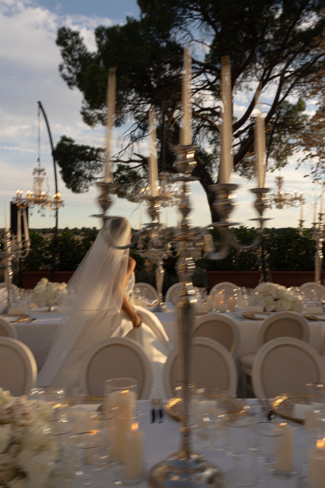 Bride in white gown walks past candlelit reception tables with tall candelabras at outdoor wedding.