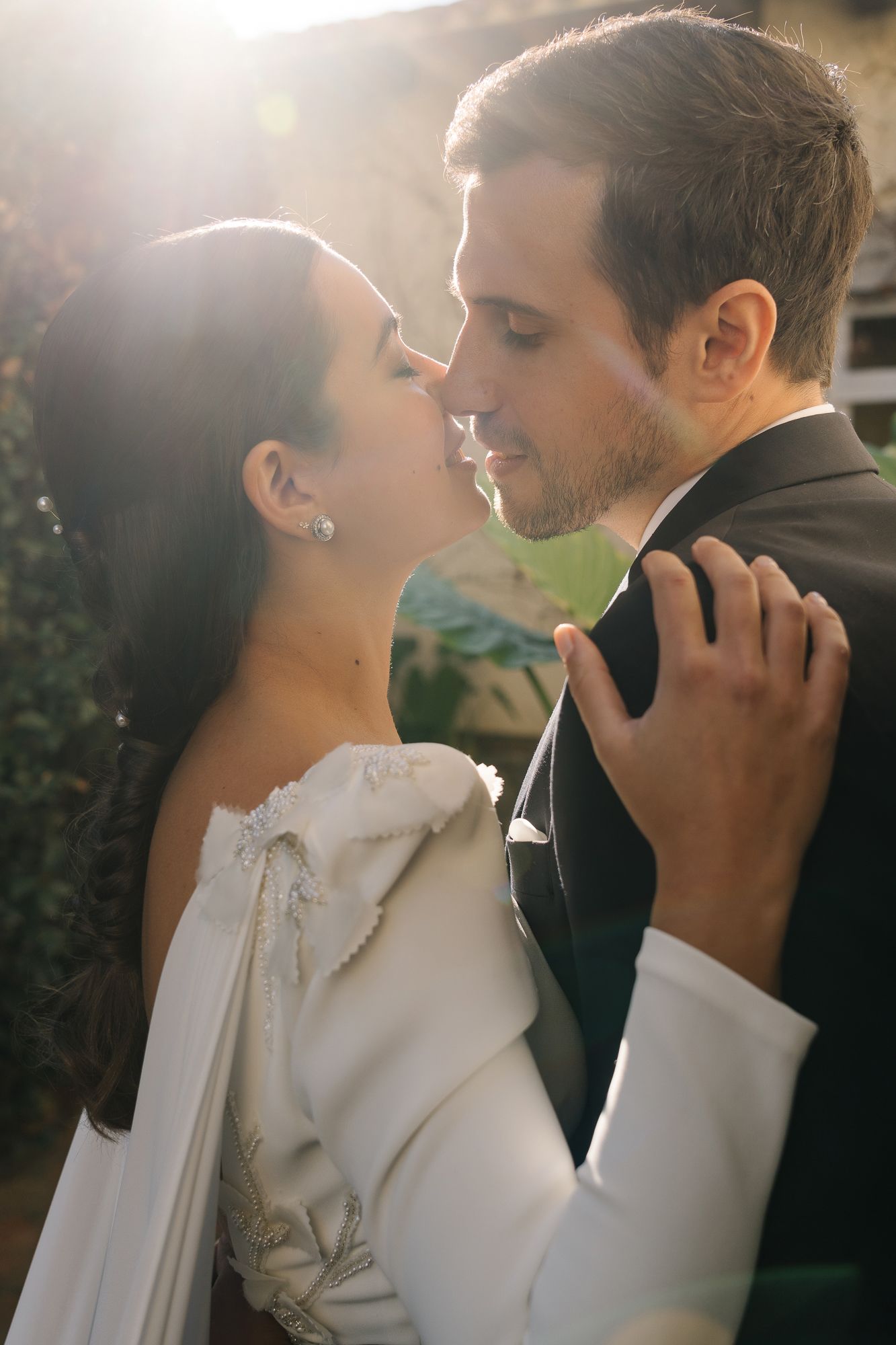 Bride and groom share a romantic almost-kiss in golden backlit sunlight, bride wearing white gown and pearl earrings.