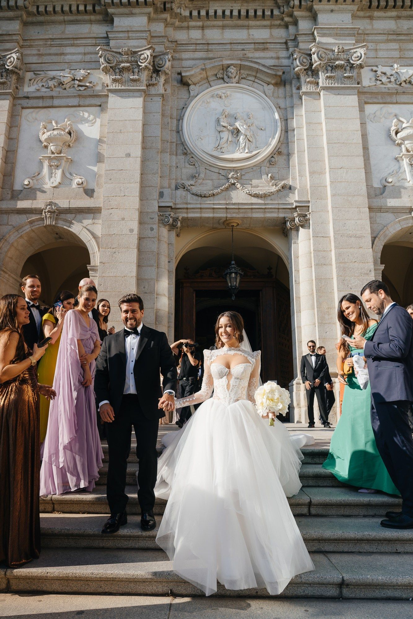 Bride in a stunning ball gown and groom in a tuxedo exit an ornate European church surrounded by wedding guests.
