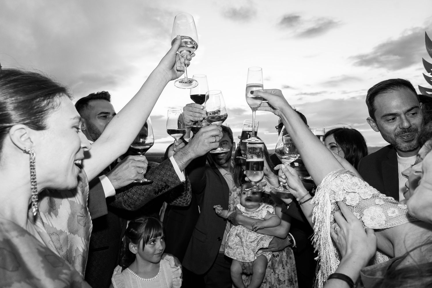 Joyful wedding guests raise glasses in a celebratory toast outdoors in a lively black and white photograph.