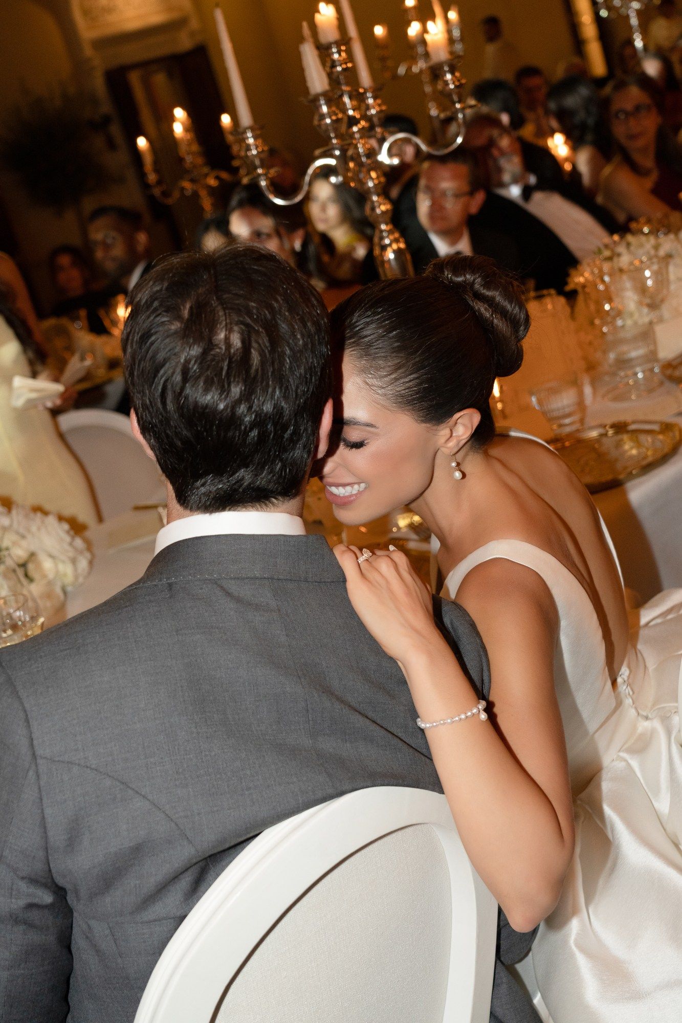 Bride in a backless satin gown smiling intimately with groom at a candlelit wedding reception dinner.