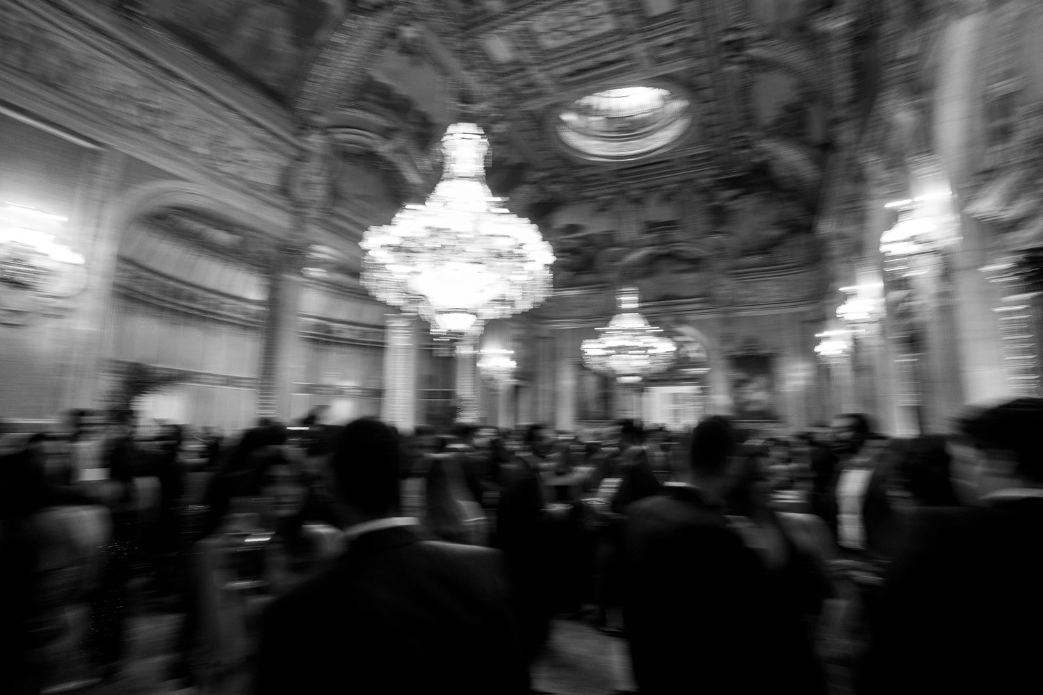 Blurred black and white photo captures elegantly dressed wedding guests mingling under grand crystal chandeliers in a ballroom.