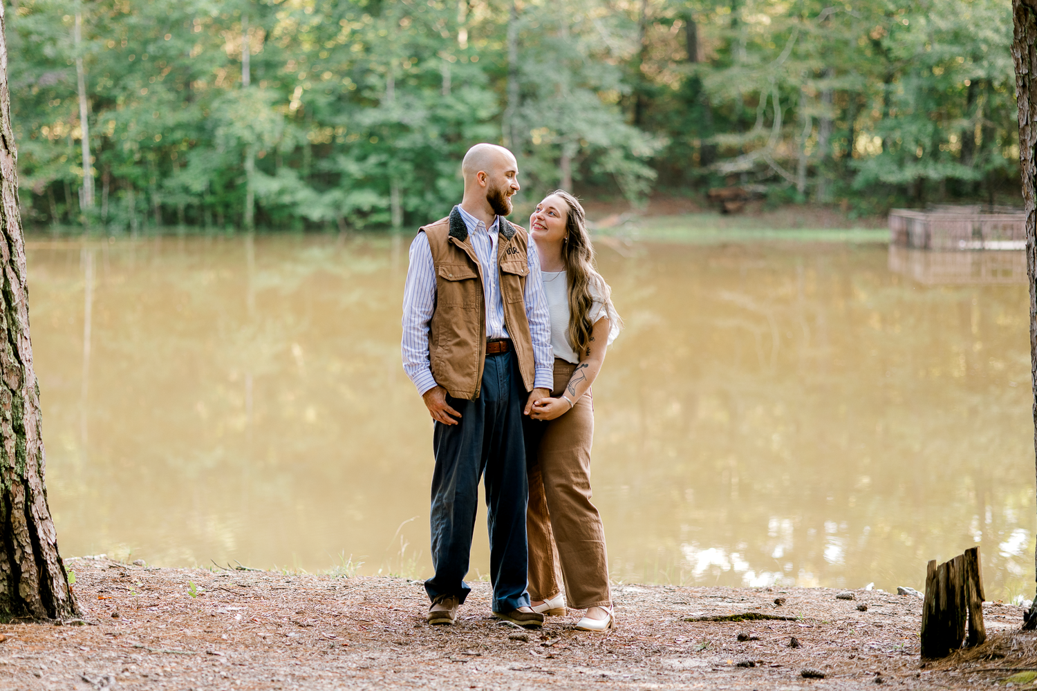 Couple in casual fall outfits embracing and smiling at each other on the shore of a calm lake at Clinton Nature Preserve near Atlanta