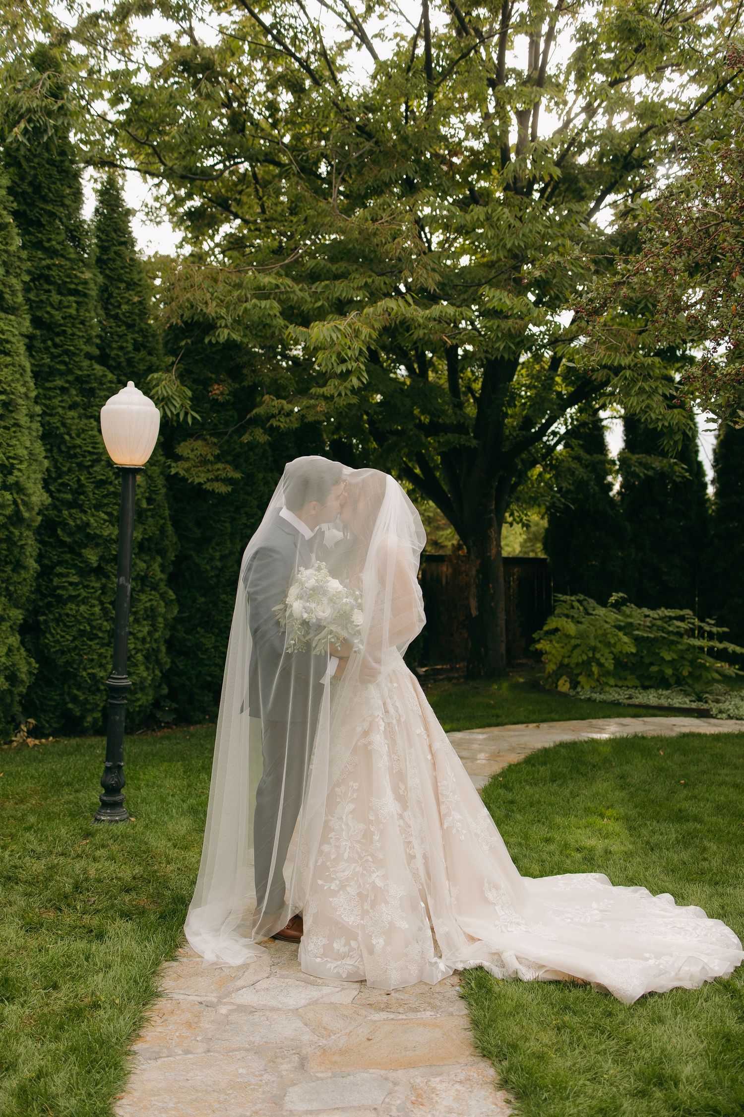 Bride and groom share a kiss under a flowing veil in a lush garden setting on their wedding day.