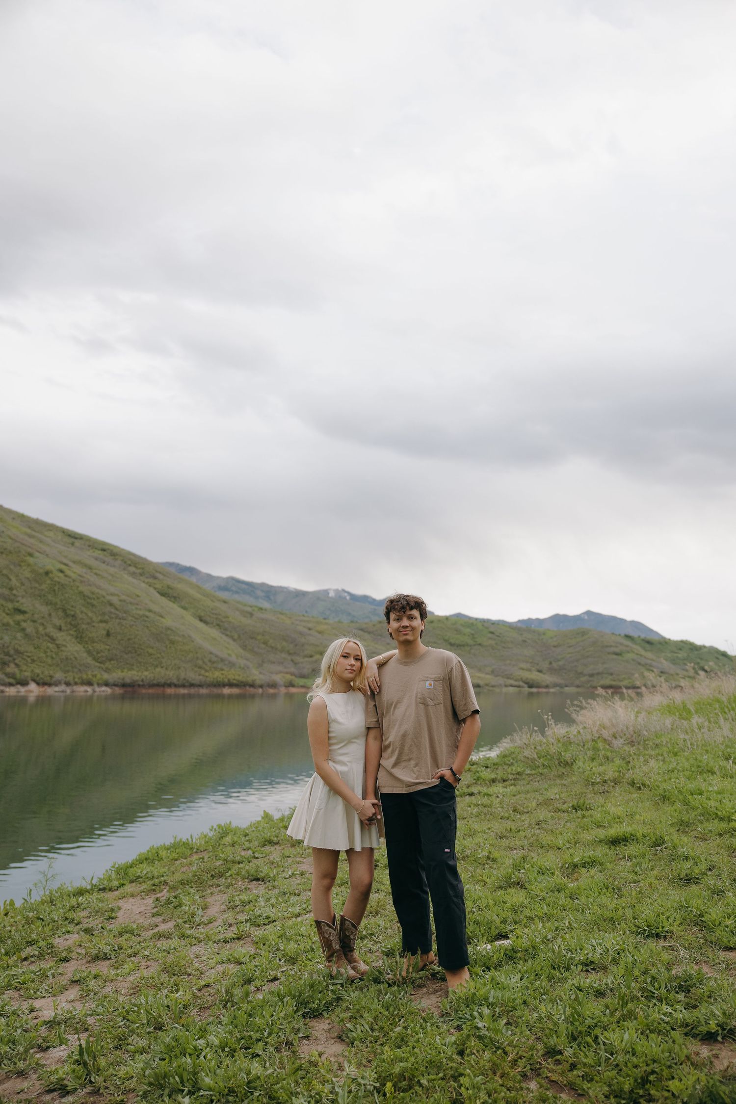 Couple holding hands standing by reflective lake with green mountains under cloudy sky.