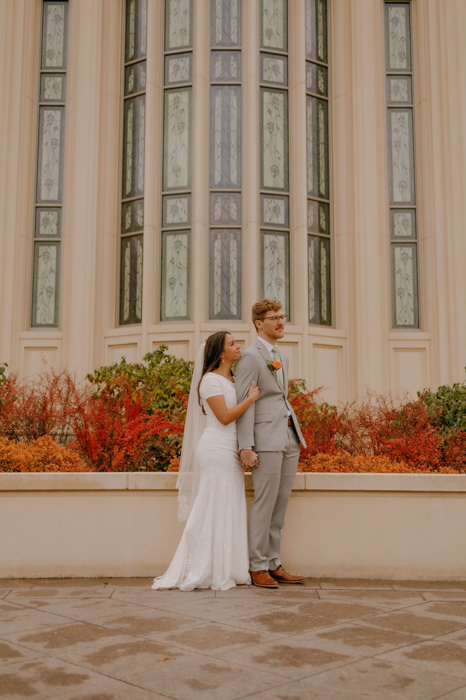 Bride in white gown and groom in gray suit standing before a grand building with autumn foliage.