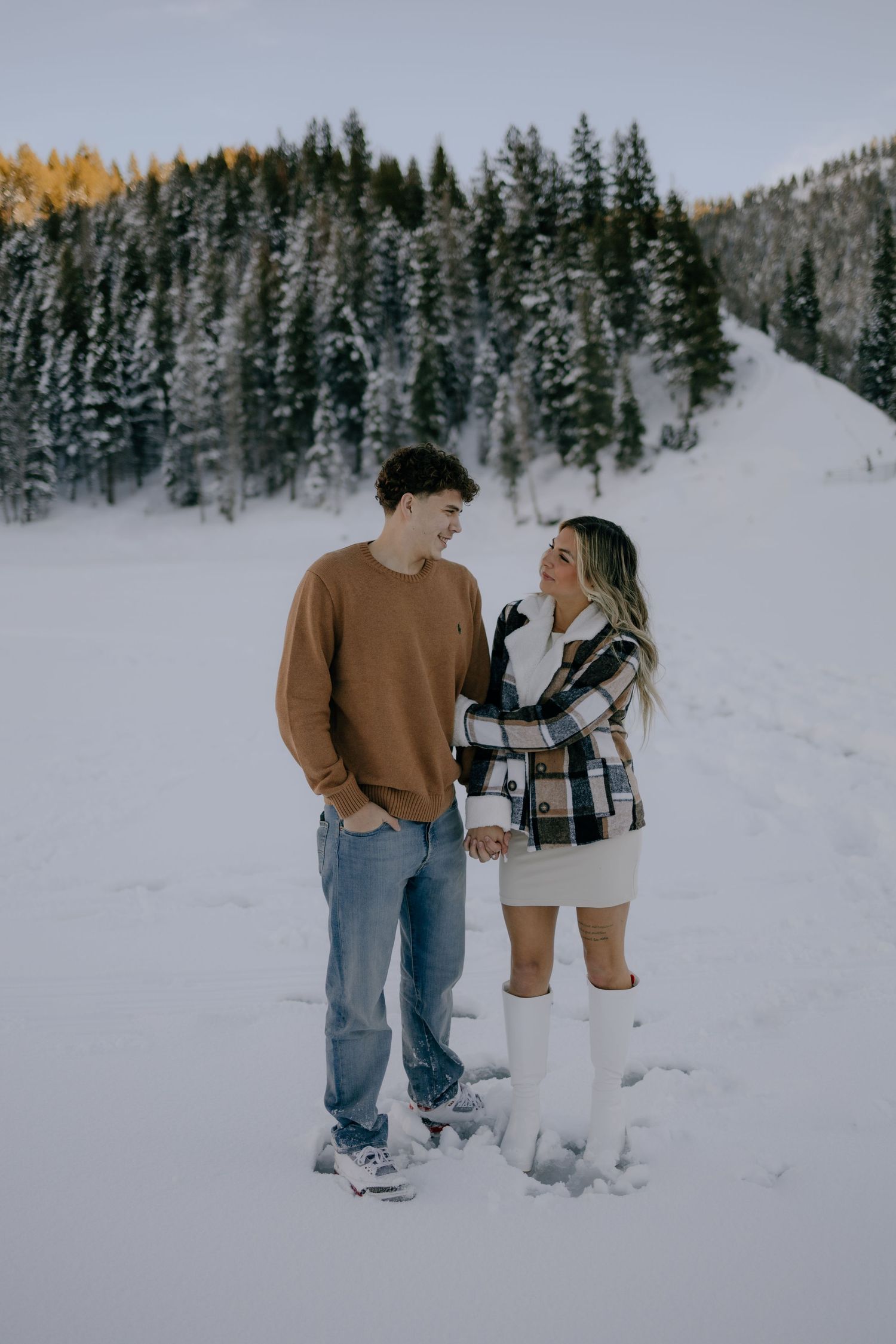 Couple standing together in a snowy mountain setting, man in brown sweater and woman in plaid jacket and white boots.