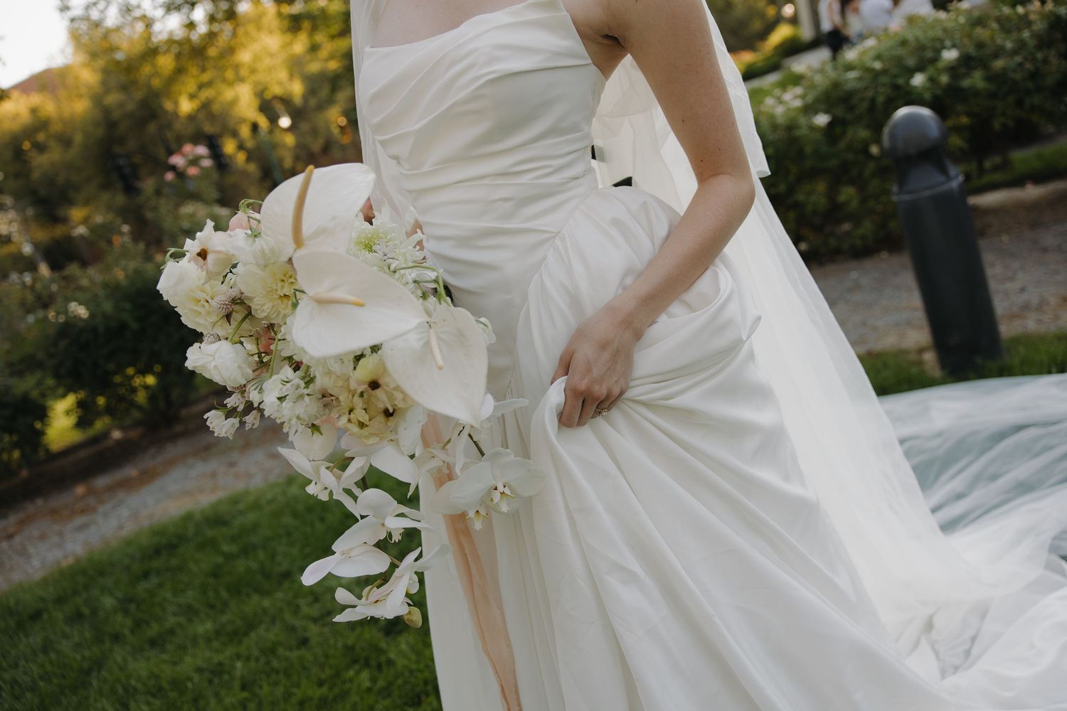 Bride in a strapless white gown holds a cascading bouquet of white calla lilies in an outdoor garden.