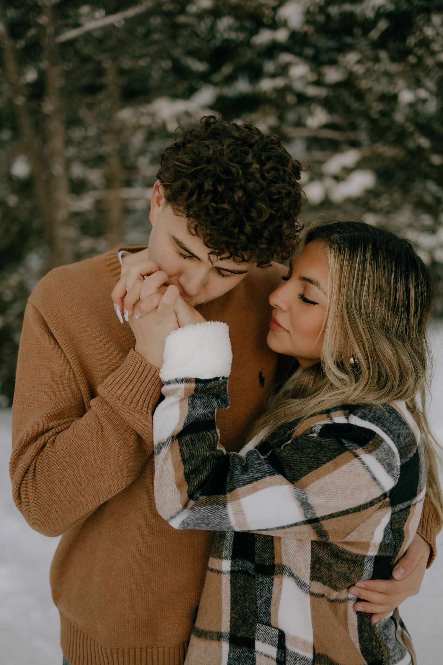 Romantic couple embracing in a snowy forest, man in tan sweater kissing woman's hands tenderly.