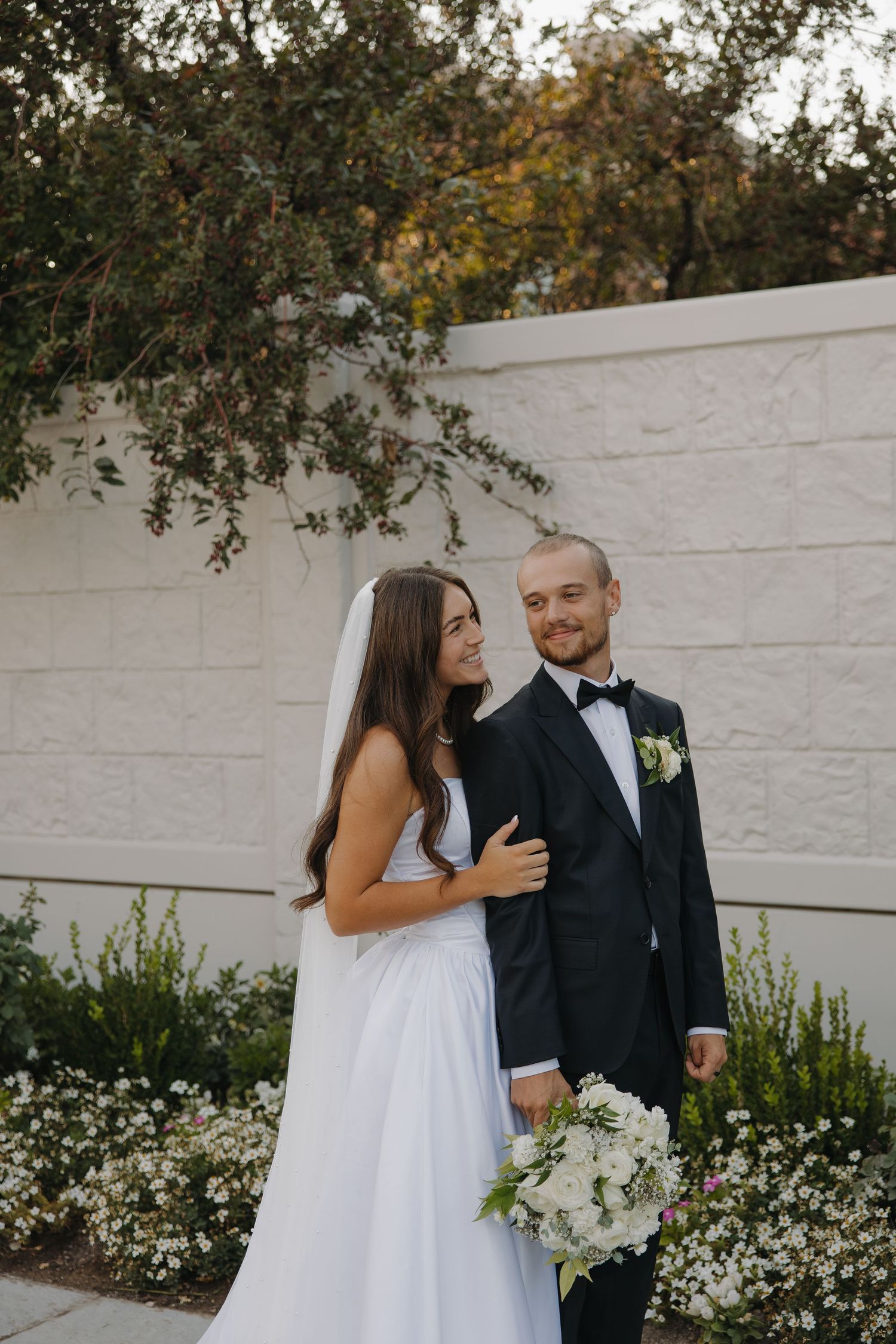 Smiling bride in white gown and groom in black tuxedo posing outdoors against a stone wall.