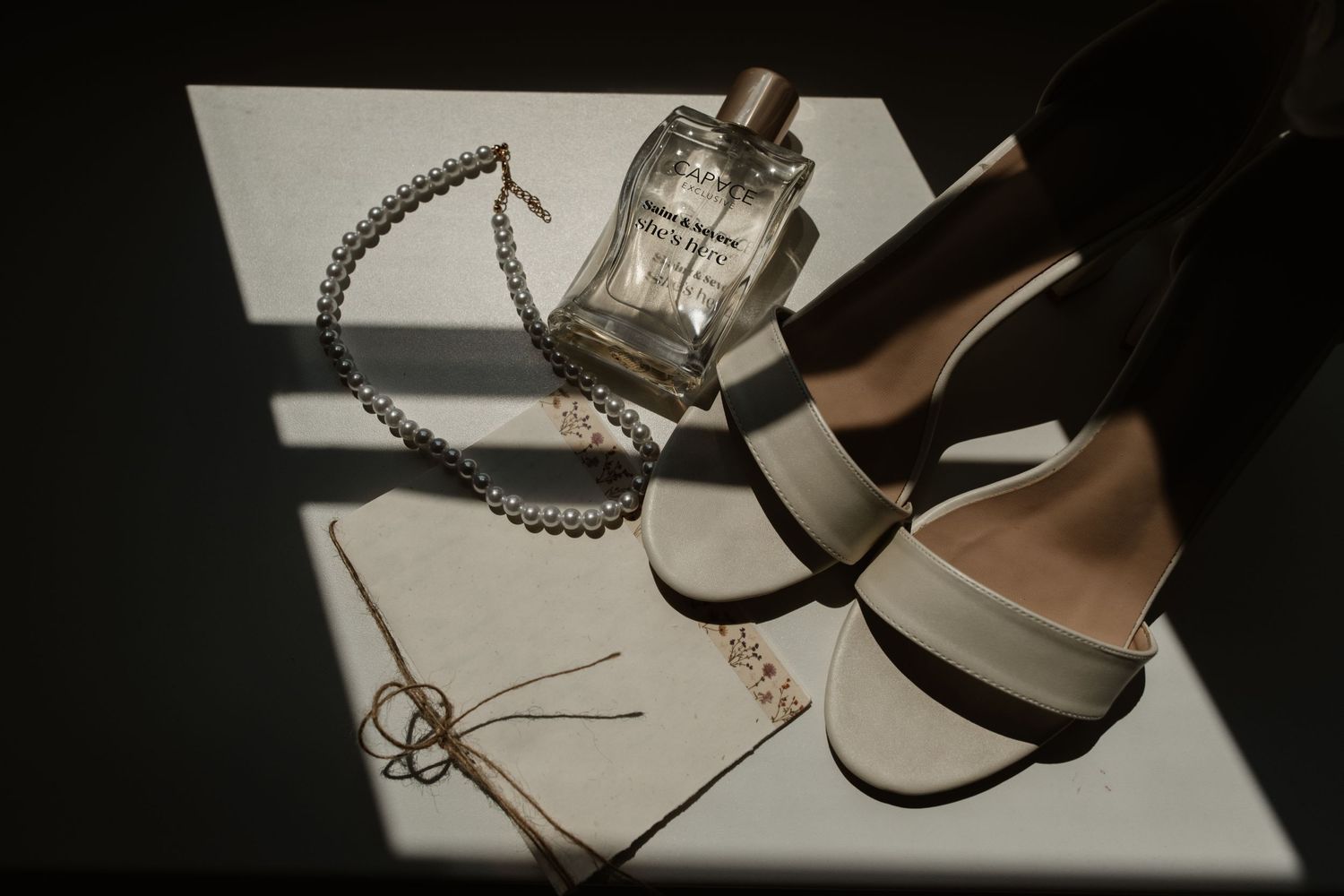 Bridal accessories flatlay featuring white heels, pearl necklace, perfume bottle, and invitation in dramatic light.