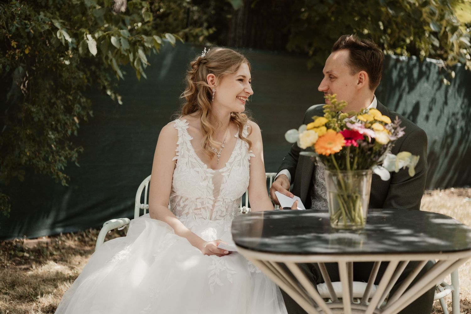 Smiling bride and groom sit together at outdoor table with vibrant floral centerpiece.