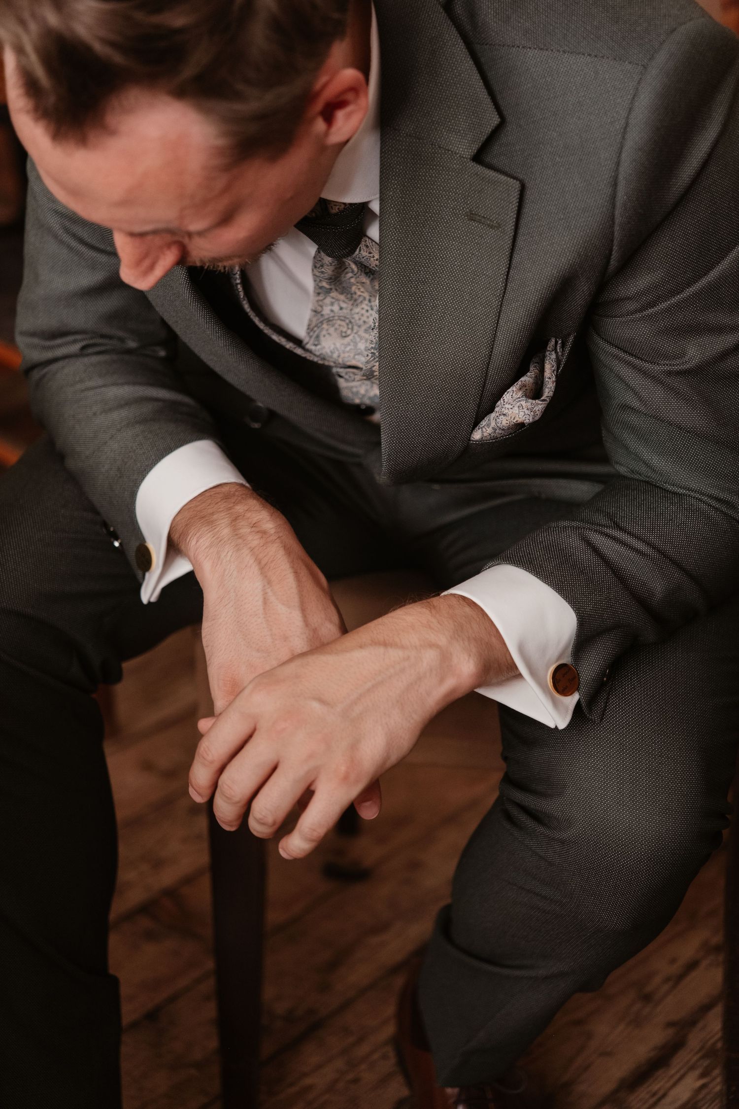 Groom in dark gray suit sits contemplatively on stool with hands clasped on wooden floor.