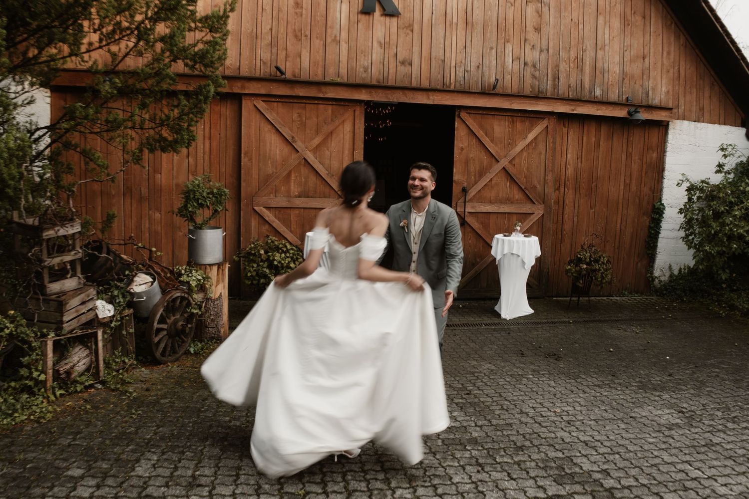 Smiling groom watches bride twirl in white wedding dress outside a rustic barn on cobblestone.