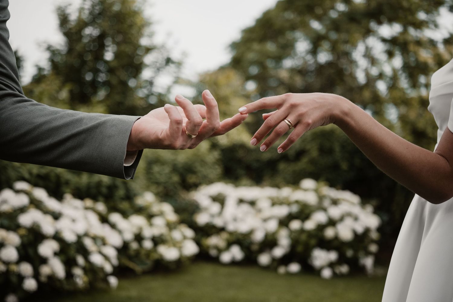 Bride and groom reach toward each other with hands near white flower garden backdrop.