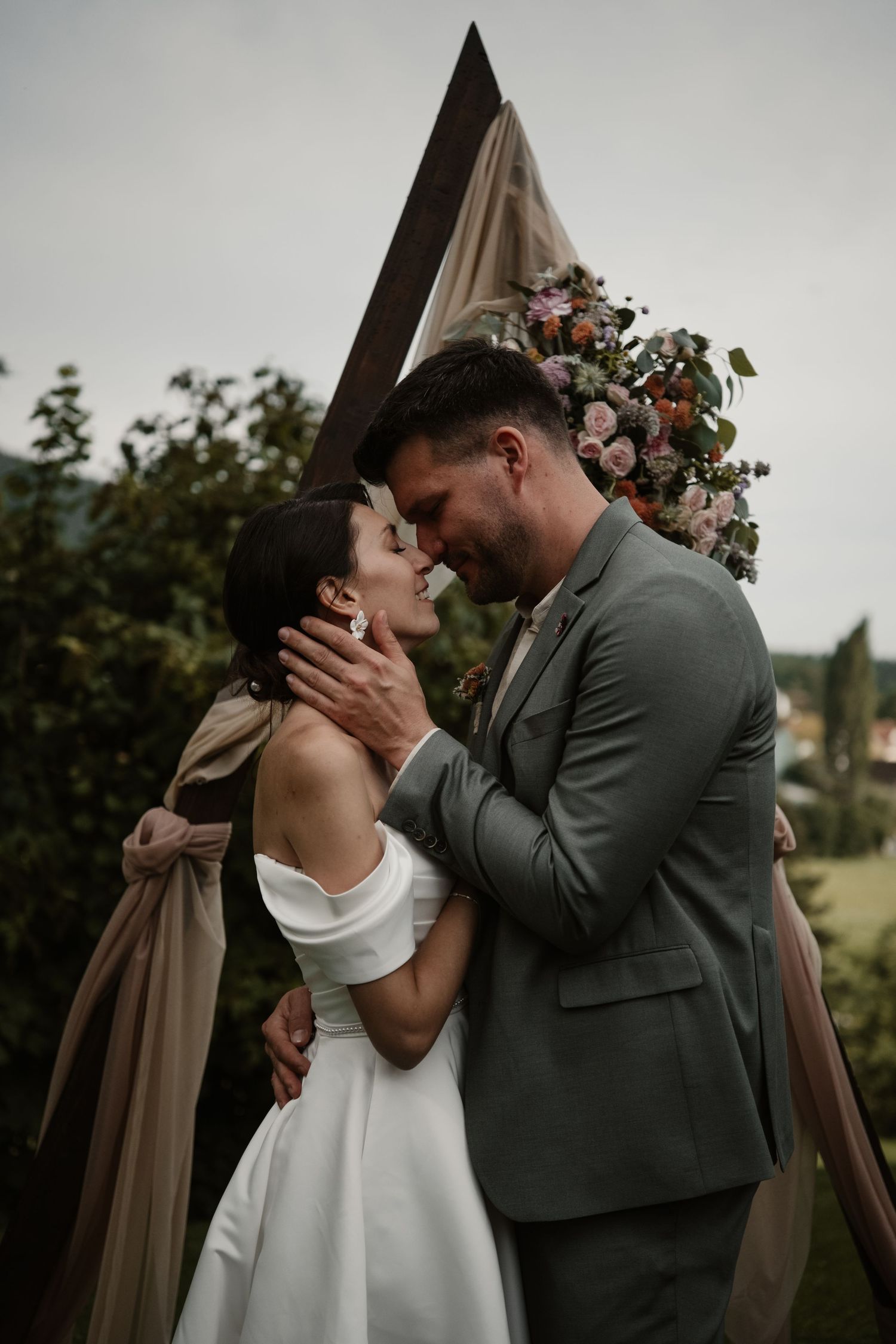 Bride and groom embrace tenderly before a triangular floral arch at outdoor wedding ceremony.