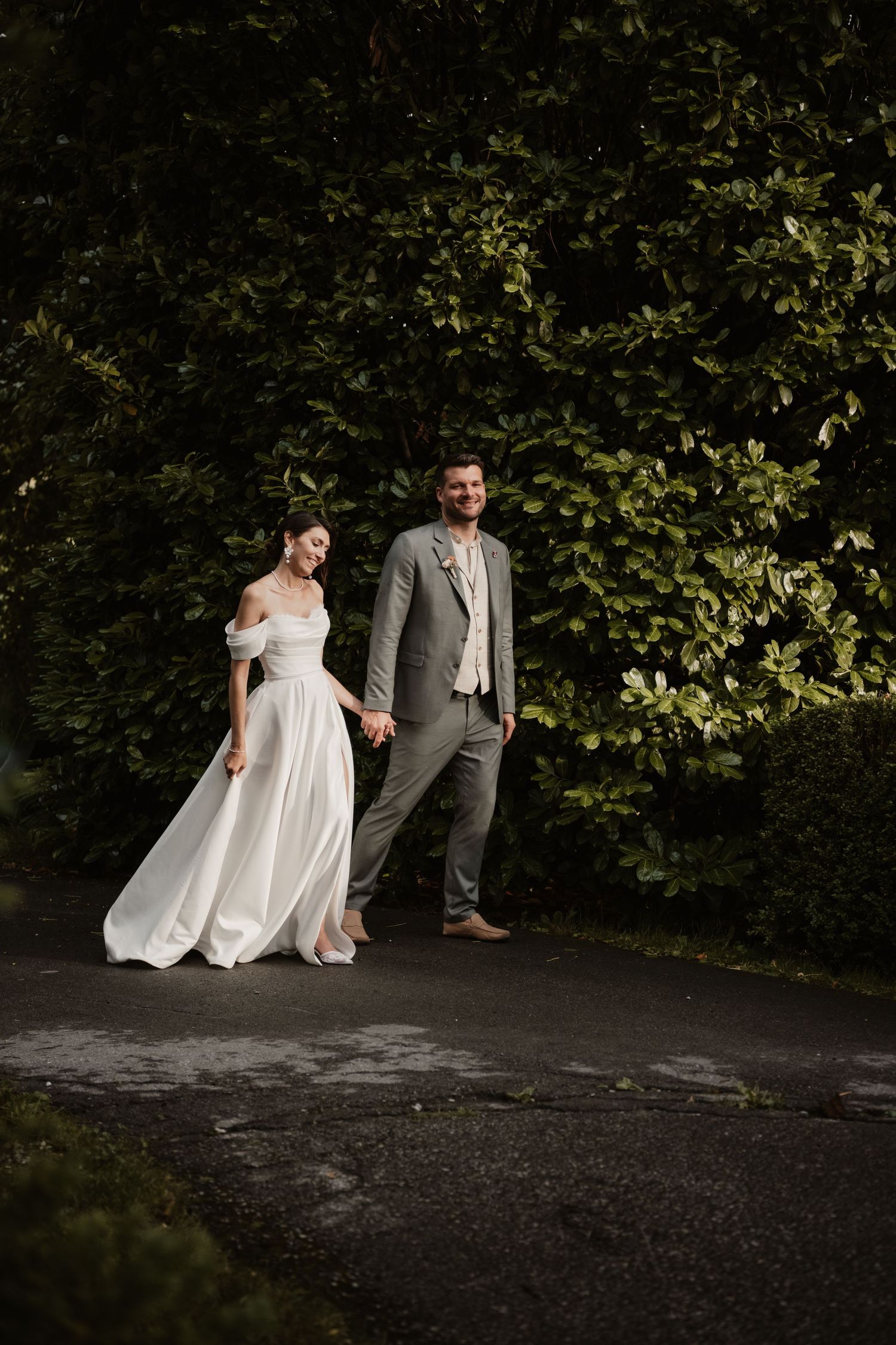 Bride and groom walk hand in hand on a shaded garden path, bride in off-shoulder white gown, groom in gray suit.