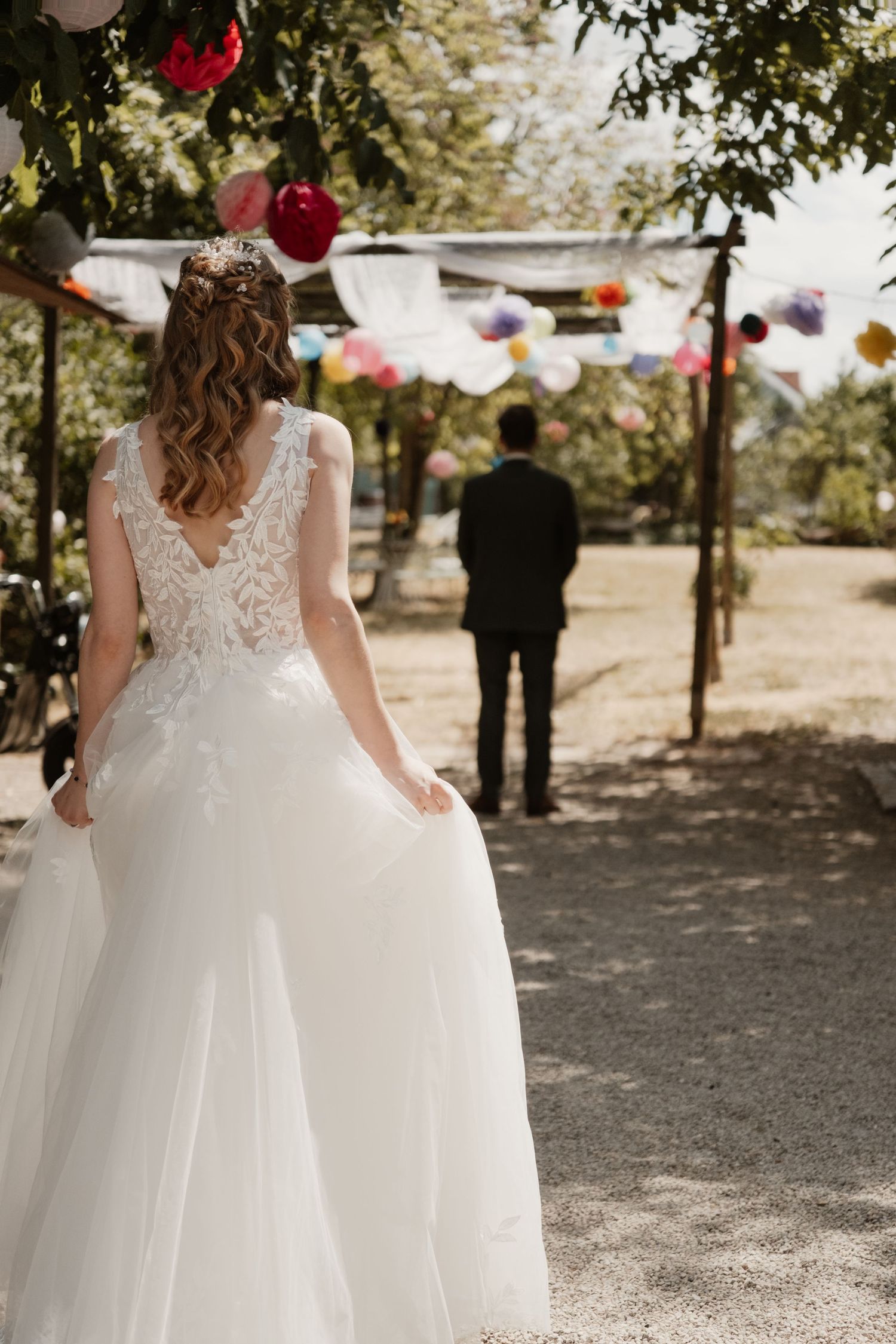 Bride in stunning lace wedding gown walks toward groom under colorful decorations at outdoor wedding.