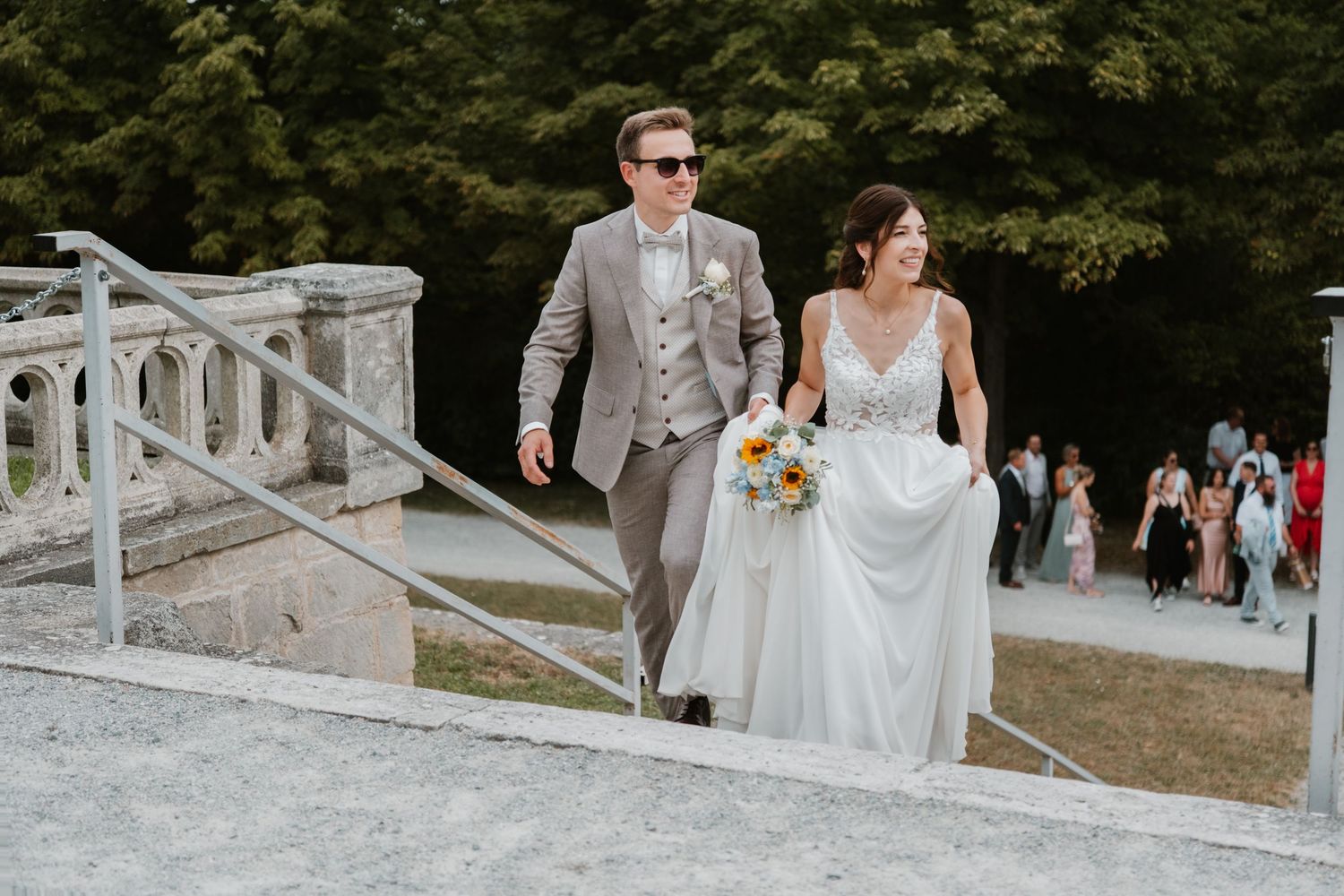 Bride and groom ascend stone stairs, bride holding colorful bouquet, guests visible in background.