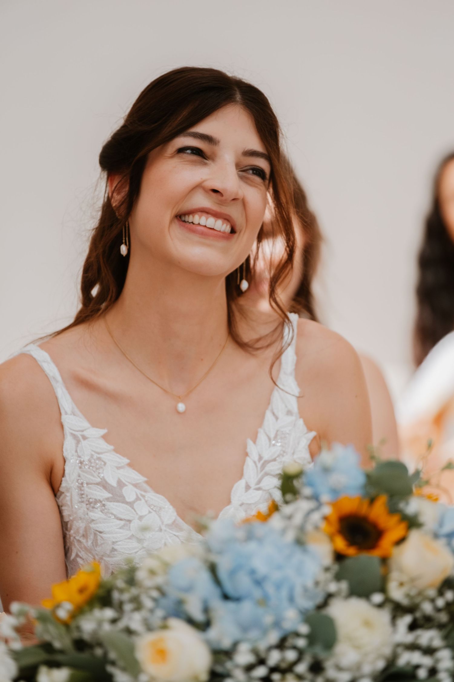 Radiant bride smiling with pearl earrings holding bouquet of sunflowers and blue hydrangeas.