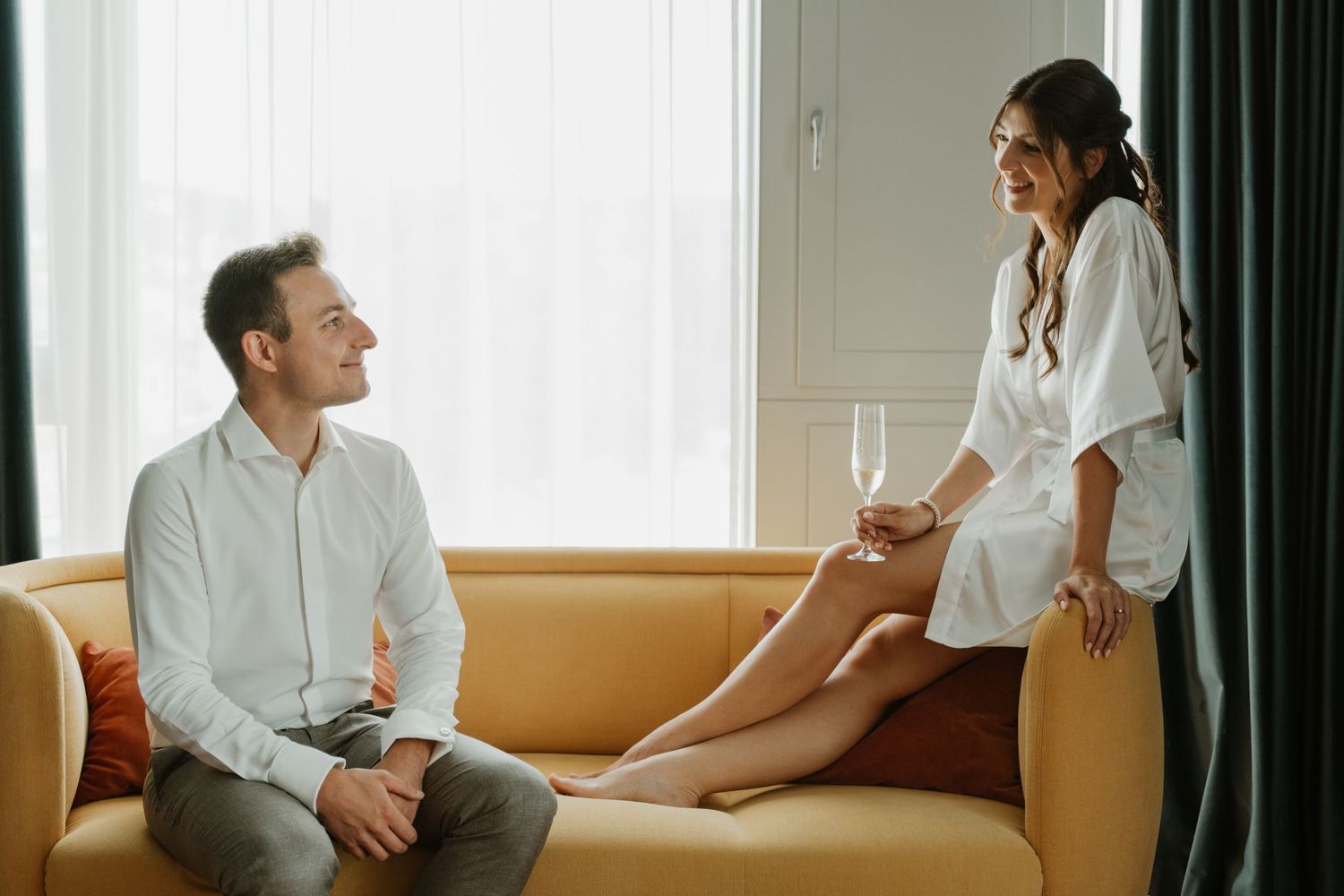 Bride in white robe holding champagne flute smiles at groom seated on yellow sofa.