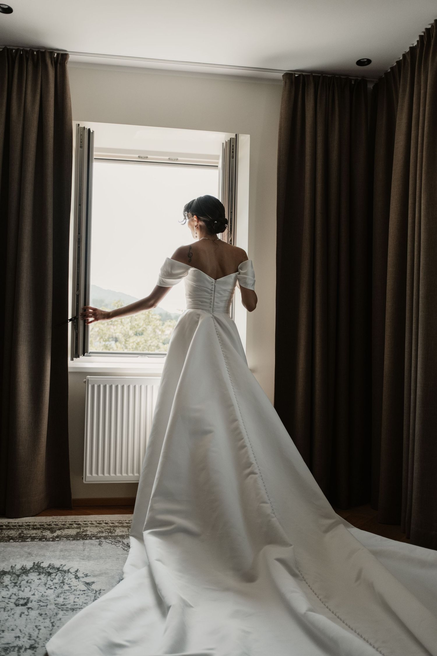 Bride in stunning off-shoulder white ball gown with long train standing at window before ceremony.