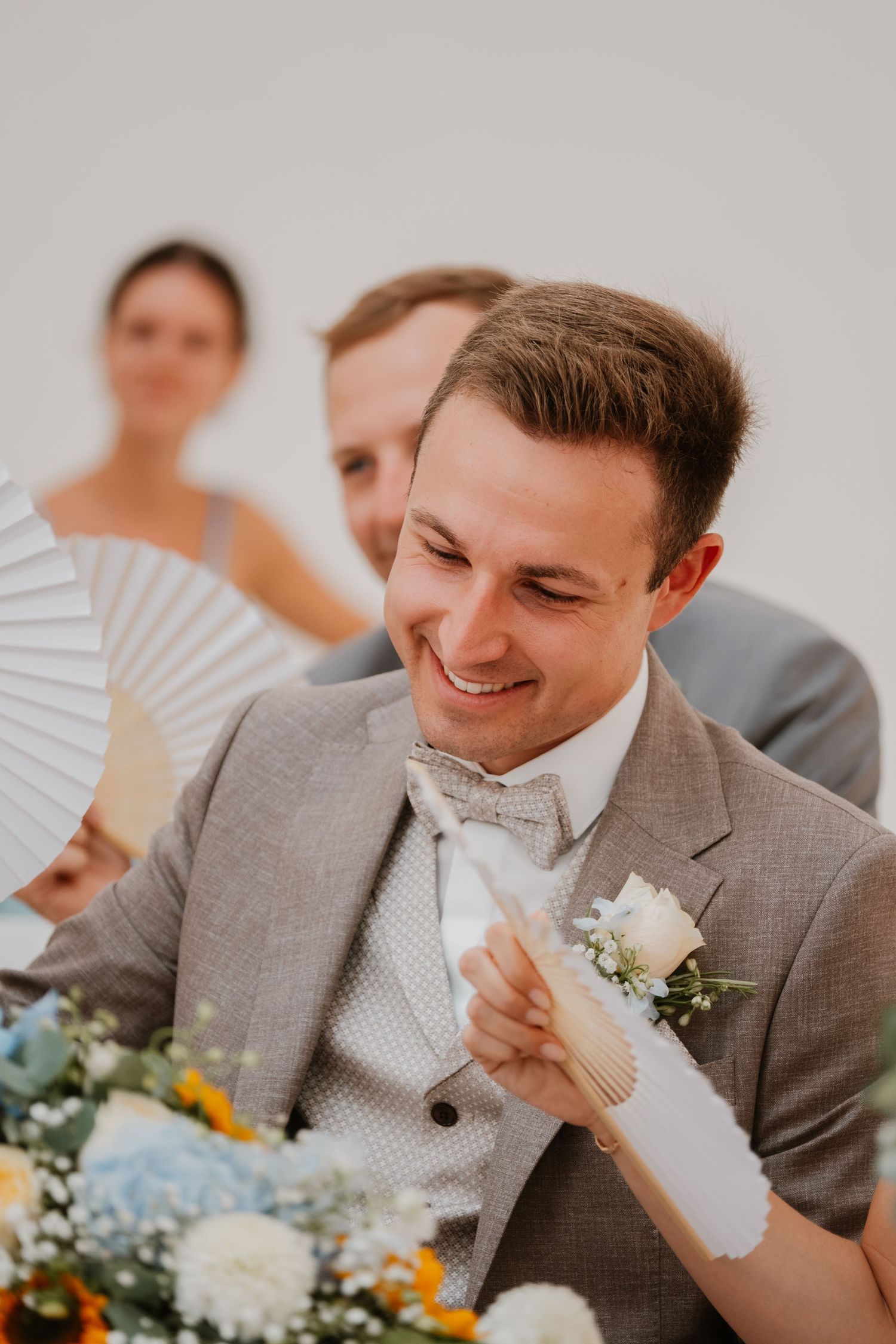 Smiling groom in gray suit with boutonniere holds a white hand fan at wedding reception.