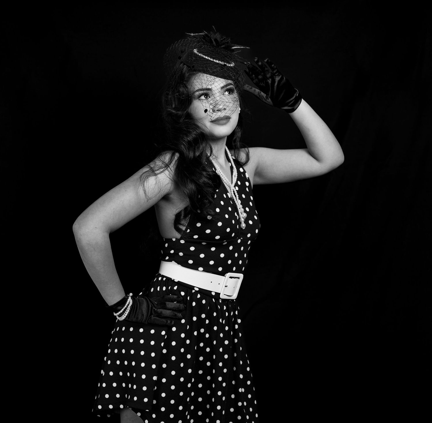 Woman in polka dot dress and hat poses confidently in black and white retro-style portrait photo.