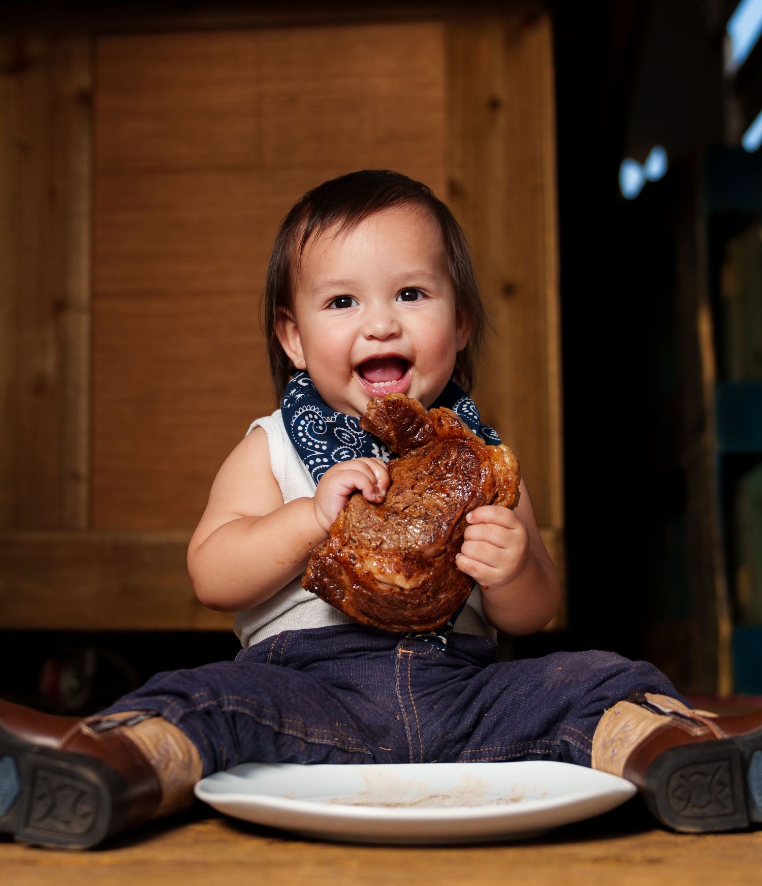 Happy baby in denim overalls and blue bandana bib eating a large piece of meat while sitting on a wooden floor.