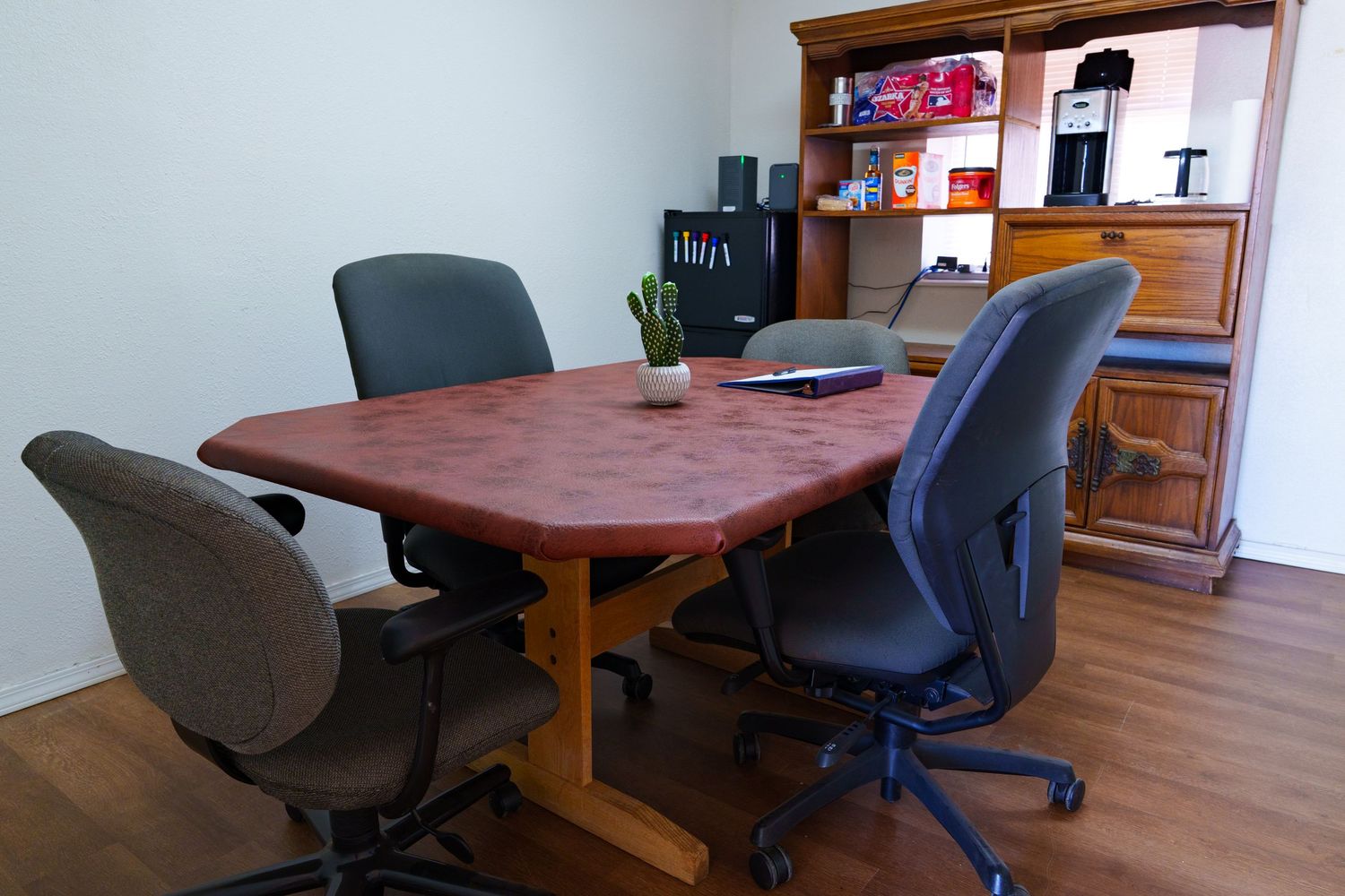 Cozy small meeting room with an oval wood table, ergonomic chairs, and a storage hutch with supplies.