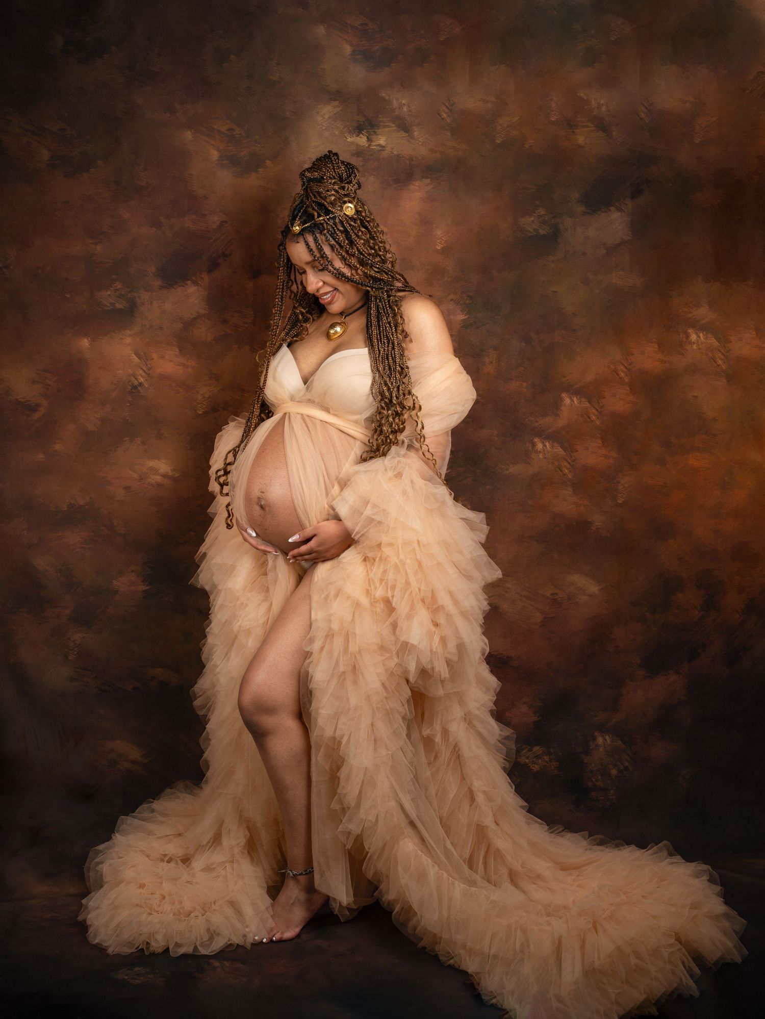 Pregnant woman in peach tulle robe with braided updo poses gracefully against a warm brown painted backdrop.