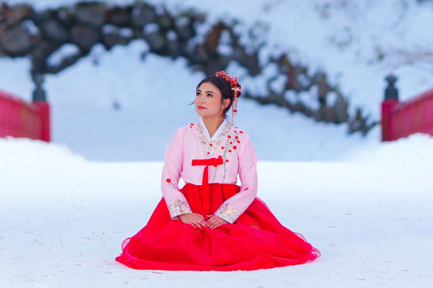 Woman in traditional Korean hanbok sits gracefully on snow, wearing red and white dress with floral hair accessories.