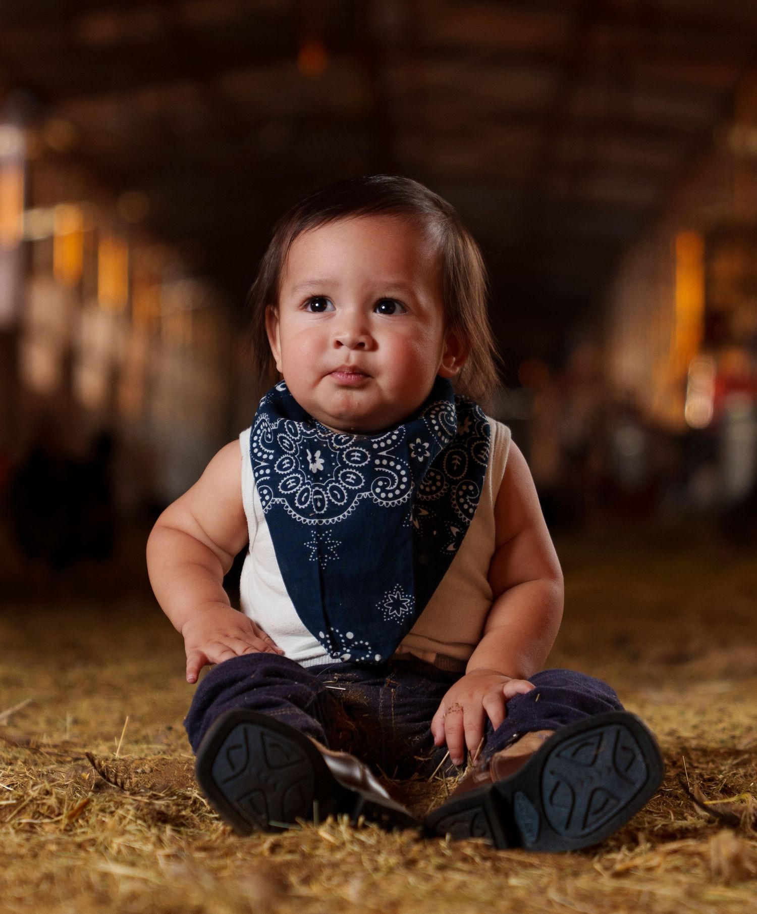 Adorable baby in navy bandana bib and jeans sits on hay in a rustic barn, styled in a cute cowboy outfit.