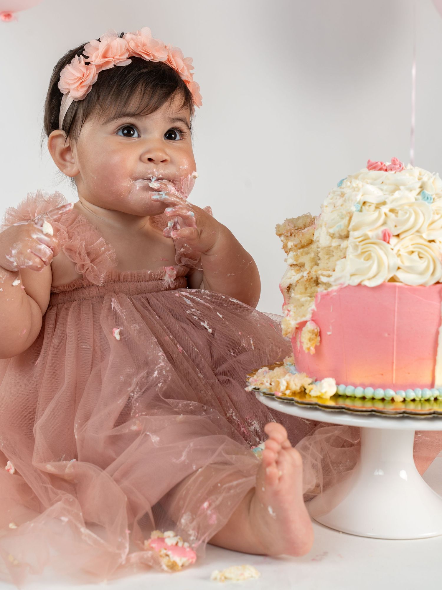 Baby girl in pink tulle dress with floral headband smashing first birthday cake on white background.