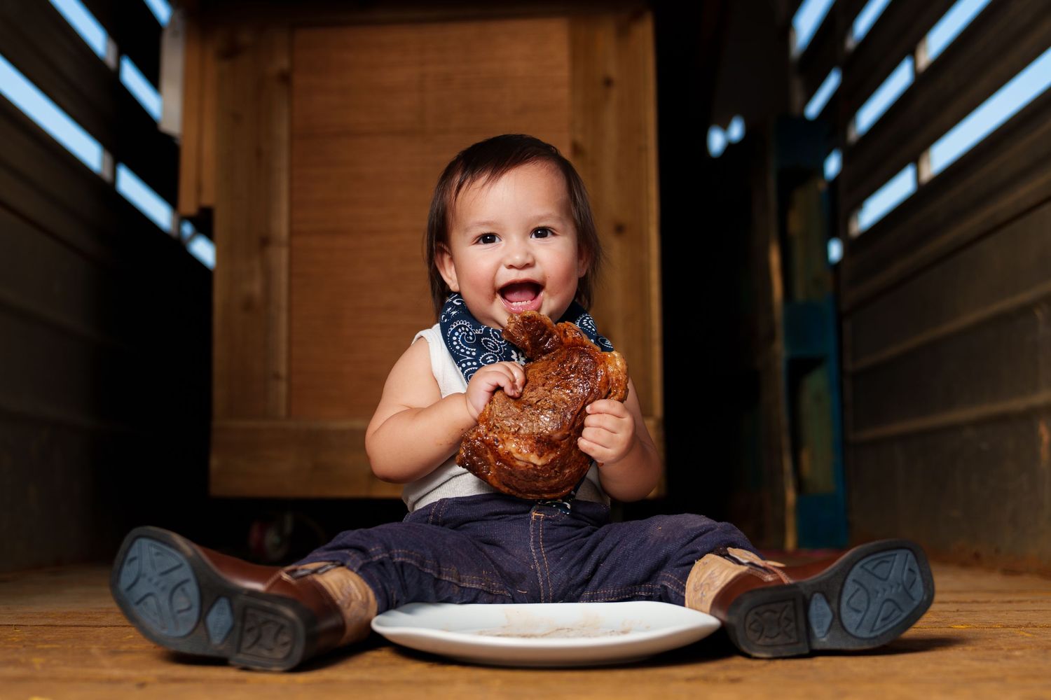 Happy baby smiling while eating a large pretzel, sitting on a wooden floor with a white plate.