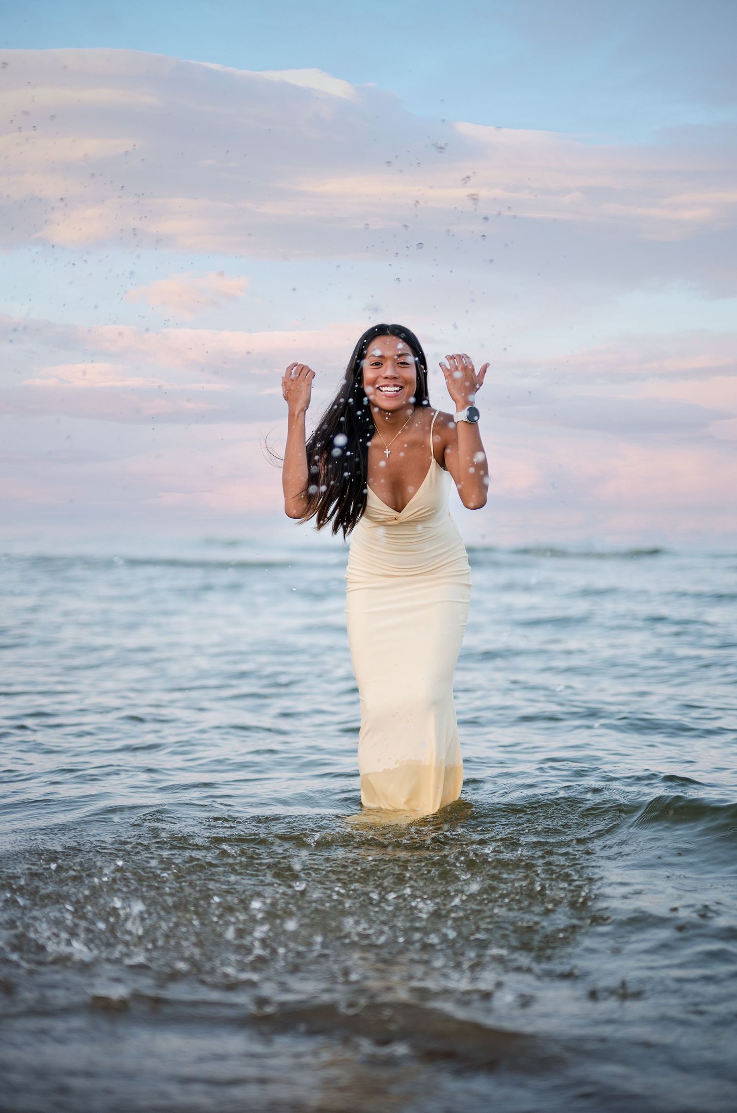 A joyful woman in a white dress stands in ocean waves at sunset, splashing water with a radiant smile.