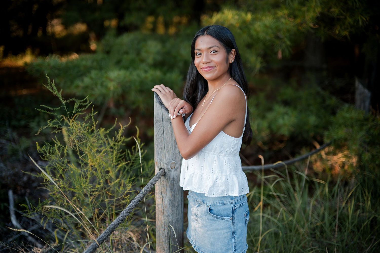 Sheboygan Senior girl in white top and denim shorts leaning on a wooden fence post in a natural outdoor setting at Kohler Andrae State Park