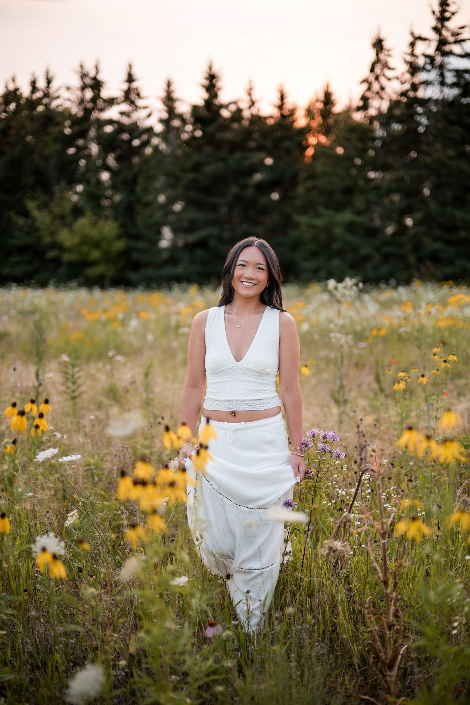 Smiling young woman in white crop top and pants walking through a wildflower field at golden hour sunset.