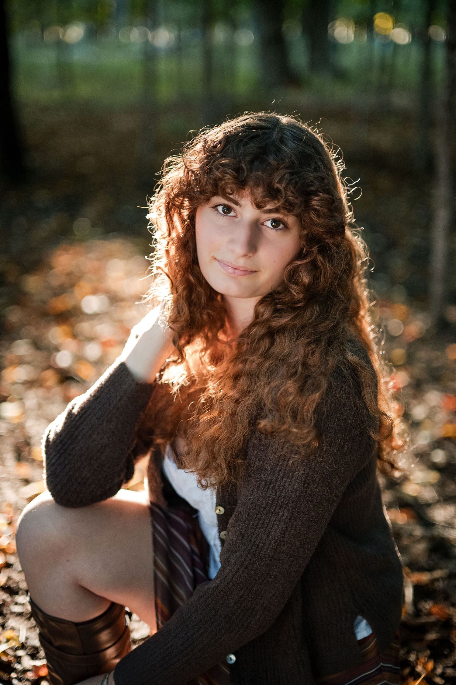 Young woman with long curly auburn hair sits smiling in a sunlit autumn forest, wearing a brown cardigan for a senior portrait.
