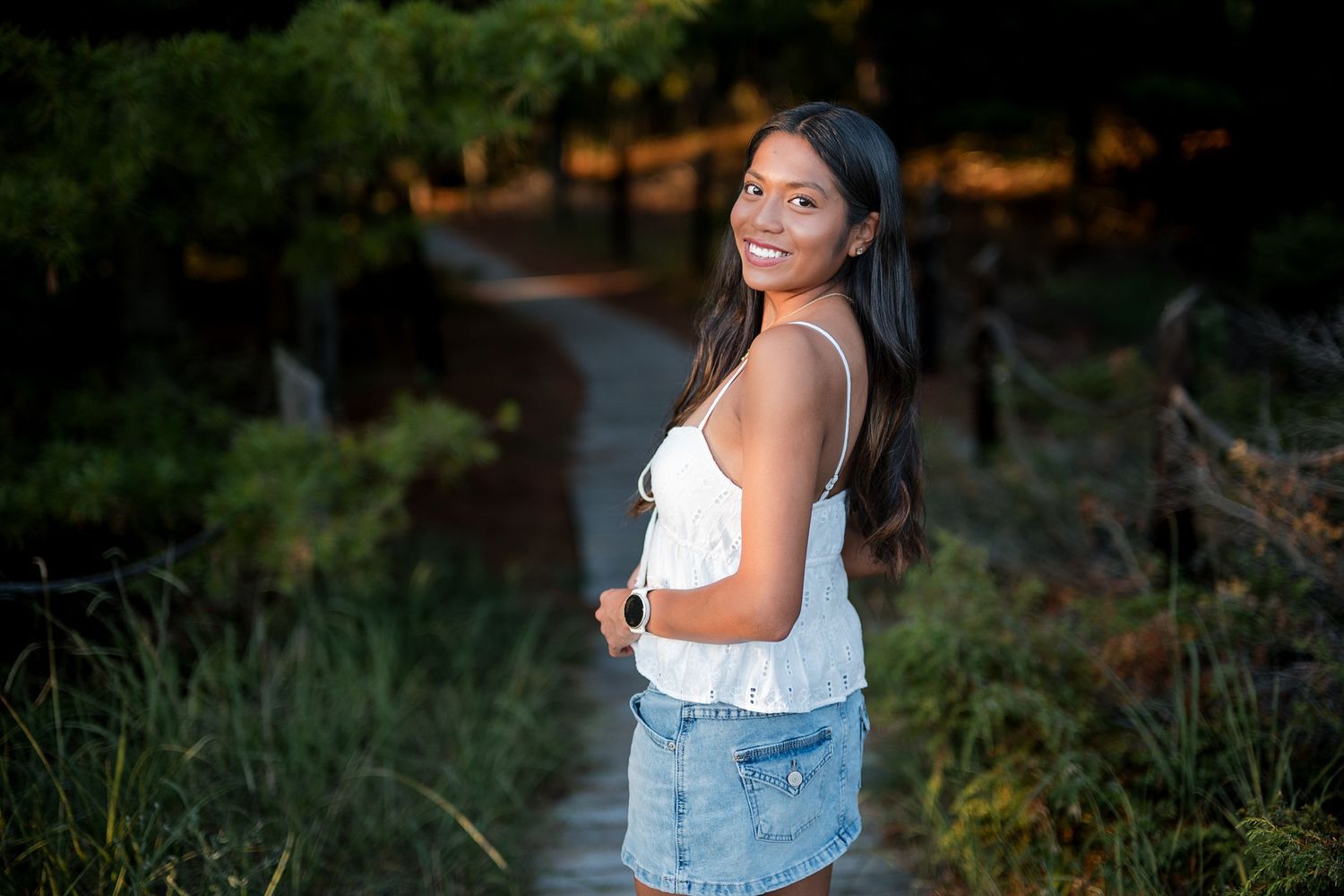 Young woman in white top and denim shorts smiling outdoors on a scenic path surrounded by lush greenery.