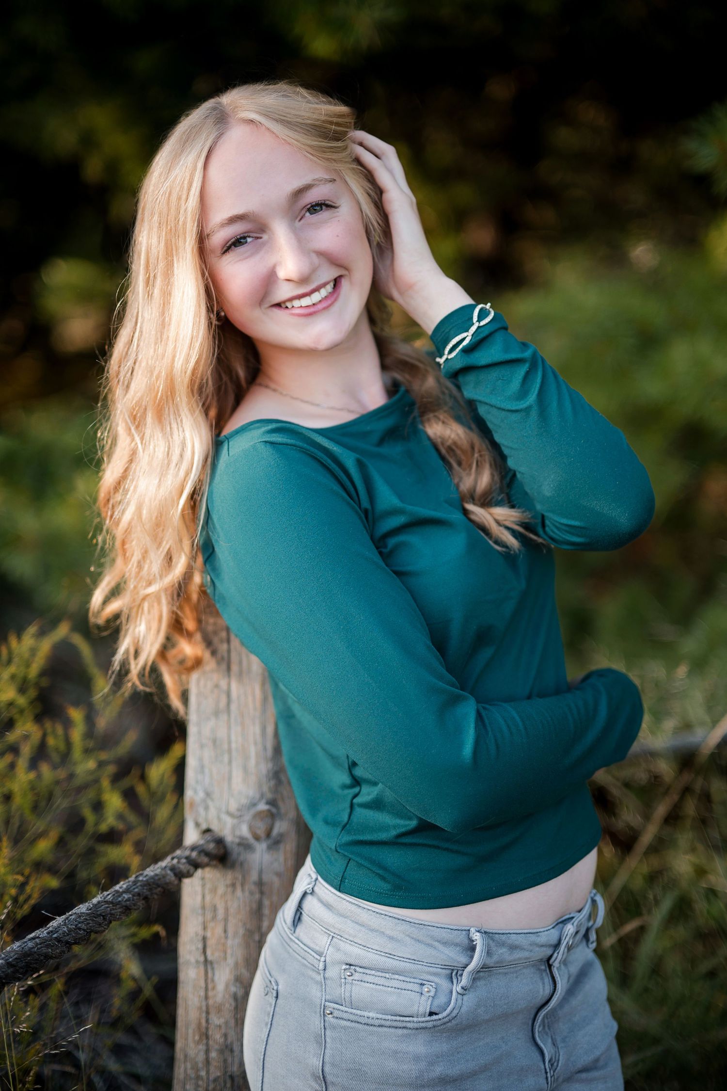 Smiling teenage girl with long blonde hair wearing a teal top and gray jeans posing outdoors near a wooden fence.