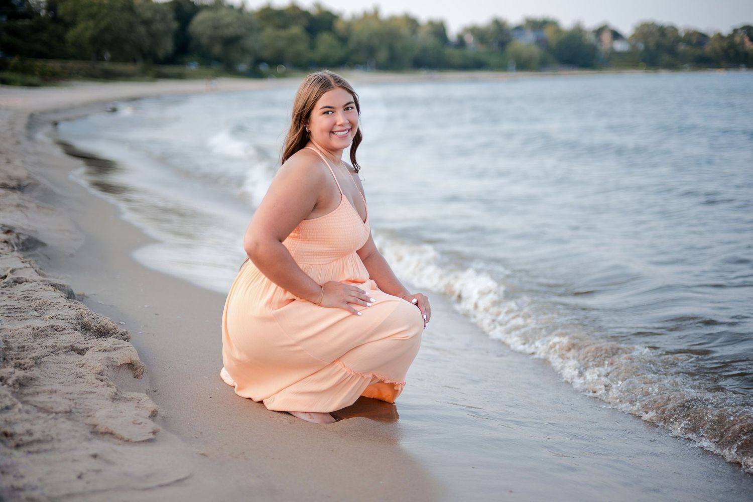 A young woman in a peach gown crouches gracefully at the shoreline of a sandy beach with gentle waves.
