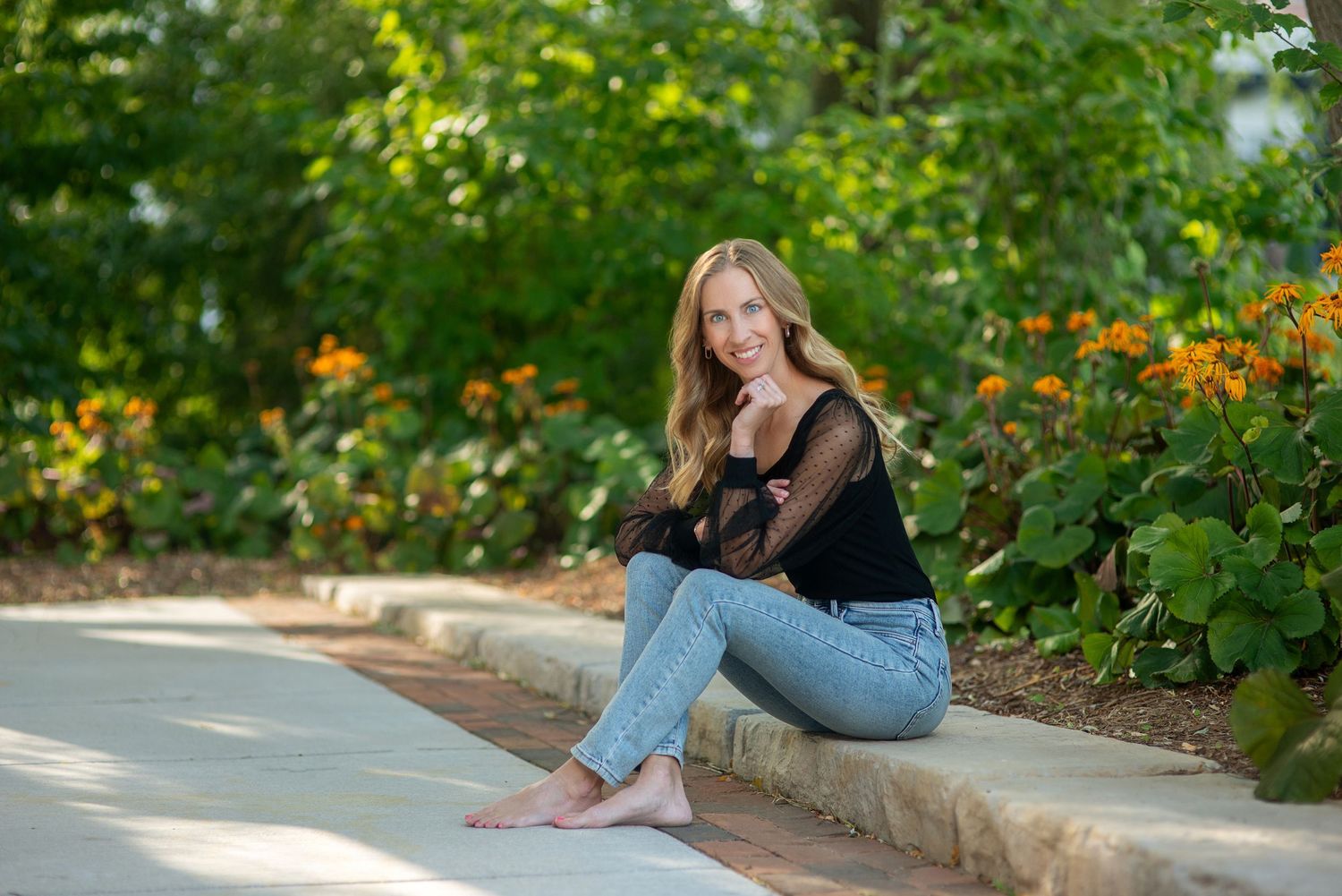 Barefoot young woman in a black top and jeans sits on a stone ledge beside orange flowers during an outdoor portrait session.